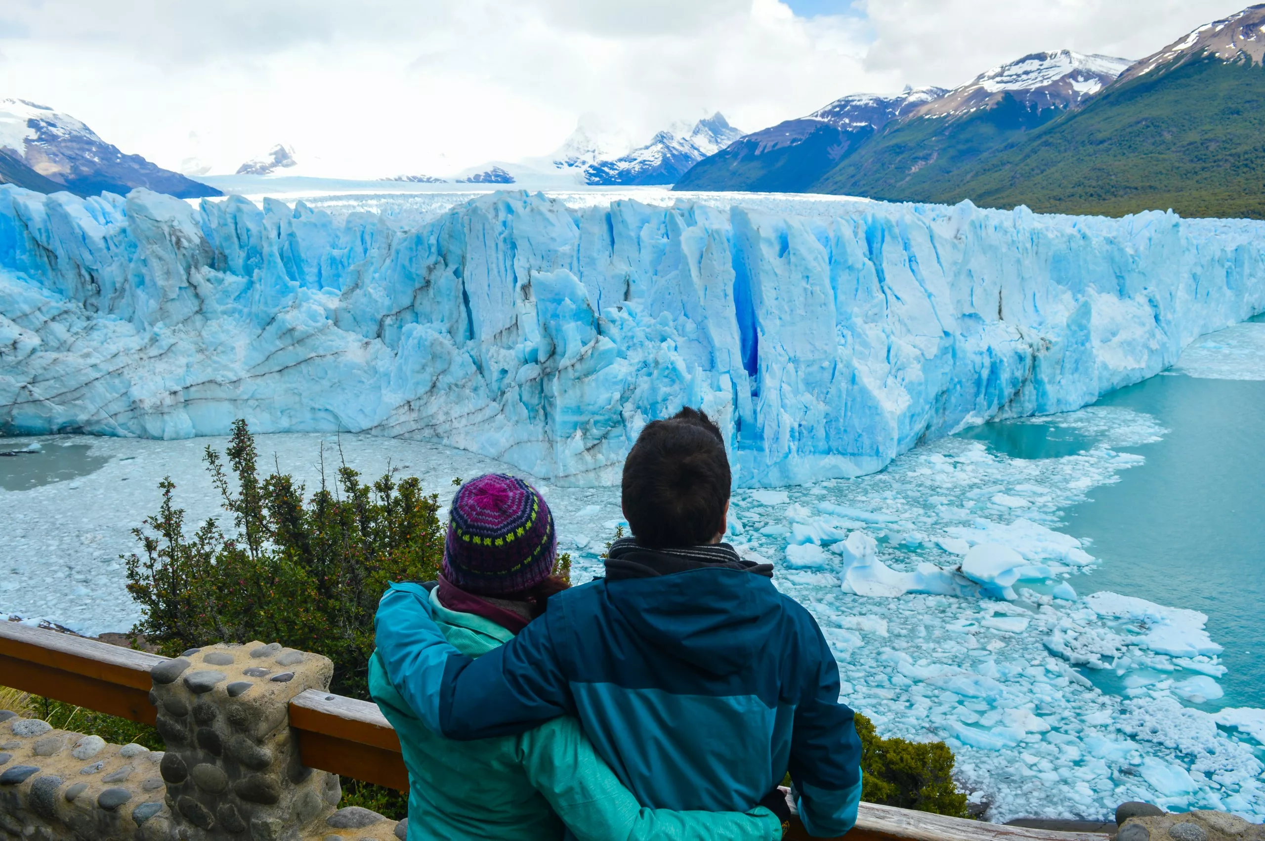Deux personnes vêtues de vestes d'hiver se tiennent sur une plate-forme d'observation en bois, contemplant le glacier Perito Moreno et des montagnes enneigées - un moment inoubliable pour ceux qui cherchent que faire en Argentine.