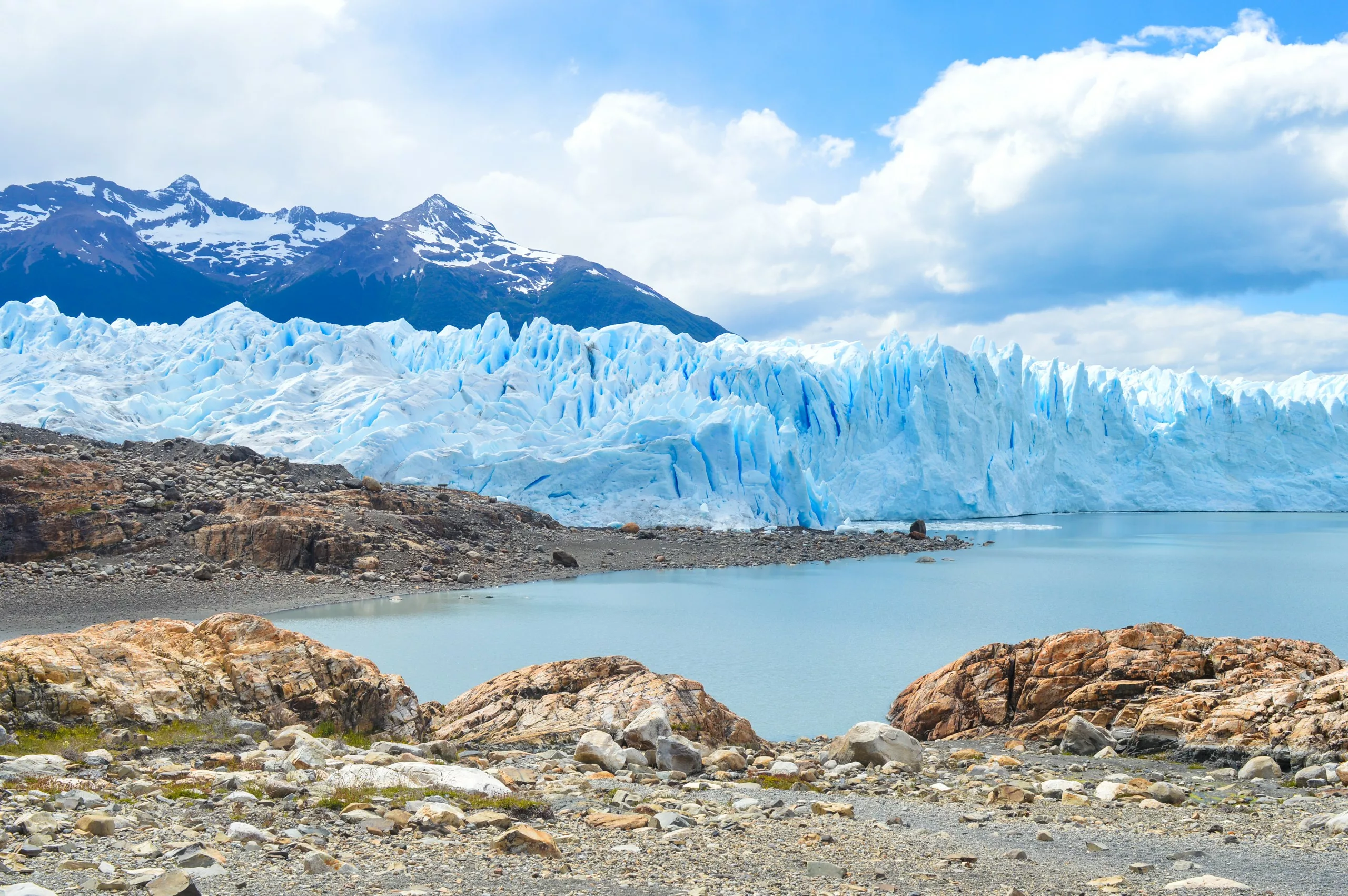 Un grand glacier avec des formations de glace déchiquetées borde un lac calme avec un terrain rocheux au premier plan. Des montagnes enneigées s'élèvent sous un ciel partiellement nuageux.