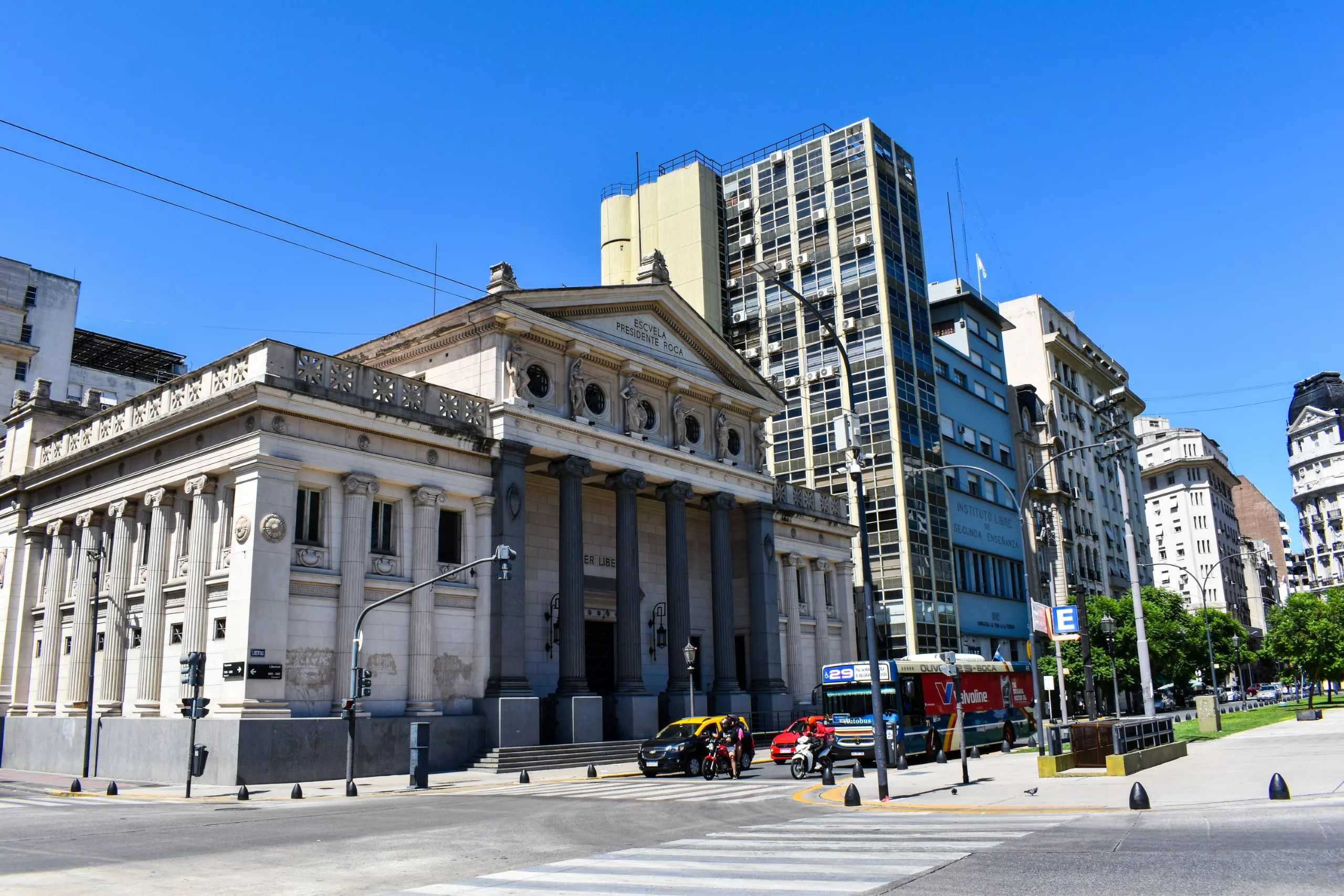 Un bâtiment néoclassique avec de hautes colonnes se dresse au coin d'une rue de Buenos Aires, entouré d'immeubles modernes et d'autres plus anciens.