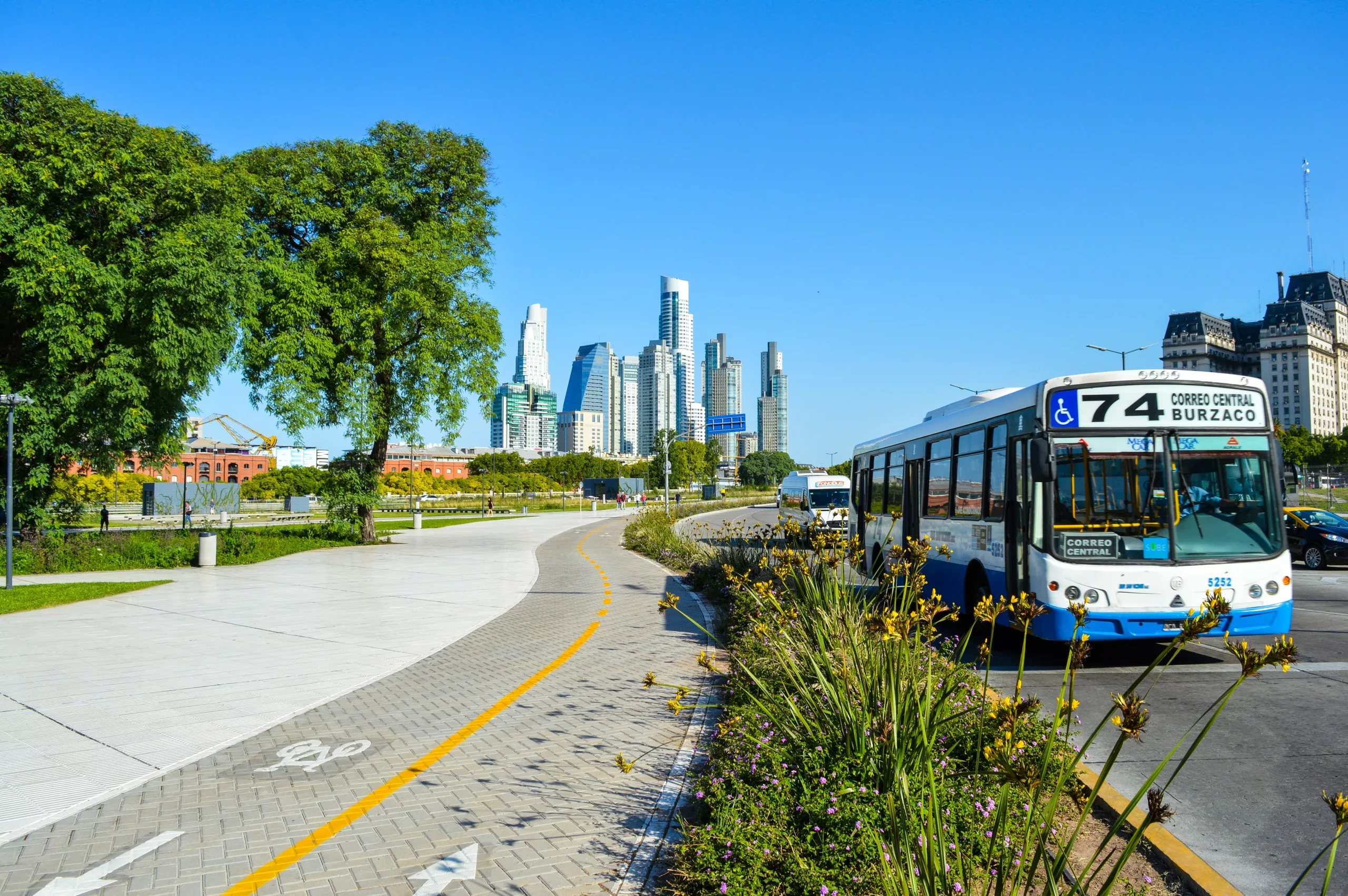 Un autobus urbain le long d'une route bordée de plantes. En arrière-plan, de grands immeubles modernes et quelques structures plus anciennes sous un ciel bleu clair.