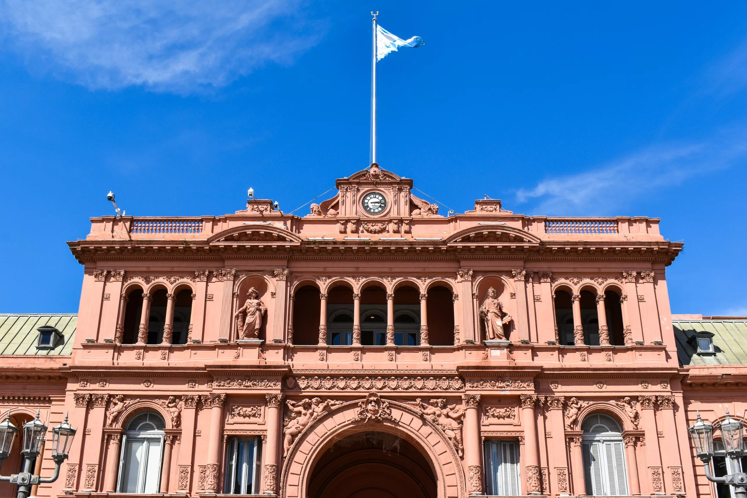 La façade d'un grand bâtiment rose avec des fenêtres cintrées, des détails ornementaux, une arche centrale et un drapeau argentin flottant au-dessus.