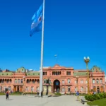 Des gens marchent devant la Casa Rosada, un grand bâtiment gouvernemental rose à visiter à Buenos Aires, en Argentine, avec le drapeau argentin flottant sous un ciel bleu clair.