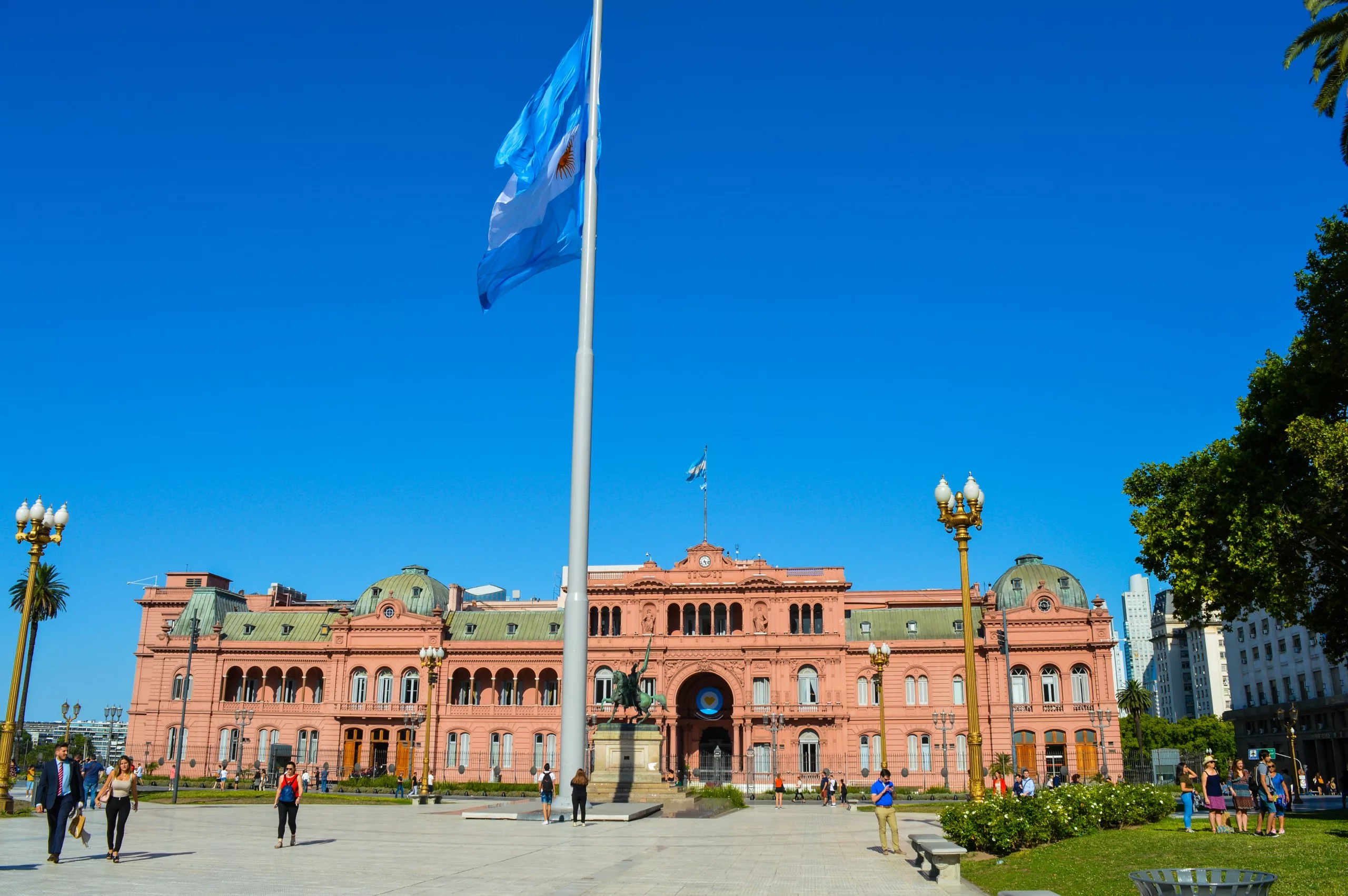 Des gens marchent devant la Casa Rosada, un grand bâtiment gouvernemental rose à visiter à Buenos Aires, en Argentine, avec le drapeau argentin flottant sous un ciel bleu clair.