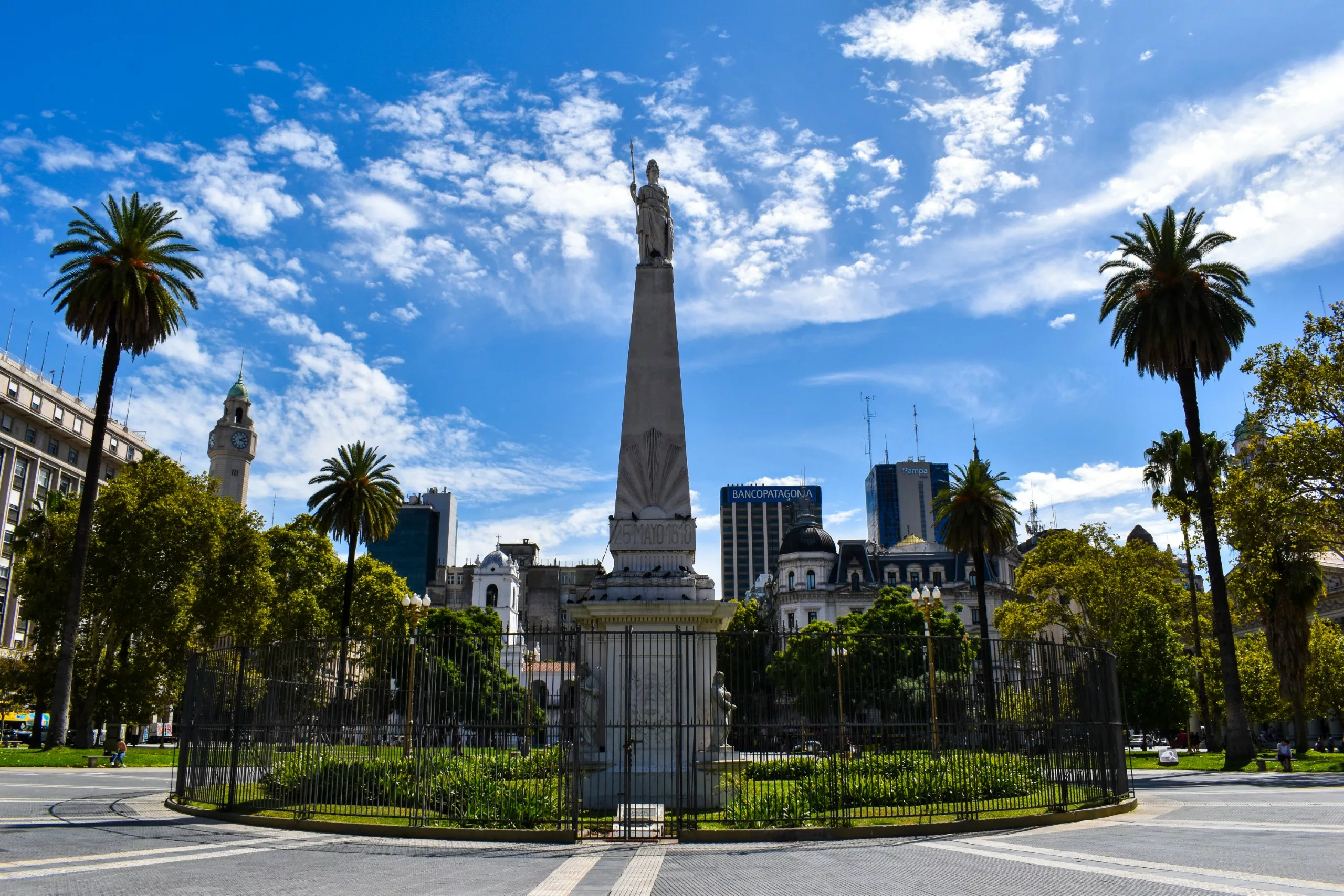 Un grand monument blanc surmonté d'une statue se dresse au centre d'une zone clôturée, entourée de verdure, de palmiers et de bâtiments historiques.