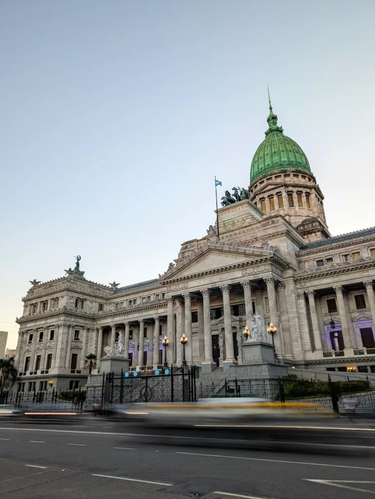 L'image montre un grand bâtiment néoclassique avec un dôme vert, des colonnes de pierre, des statues et un drapeau argentin au sommet.