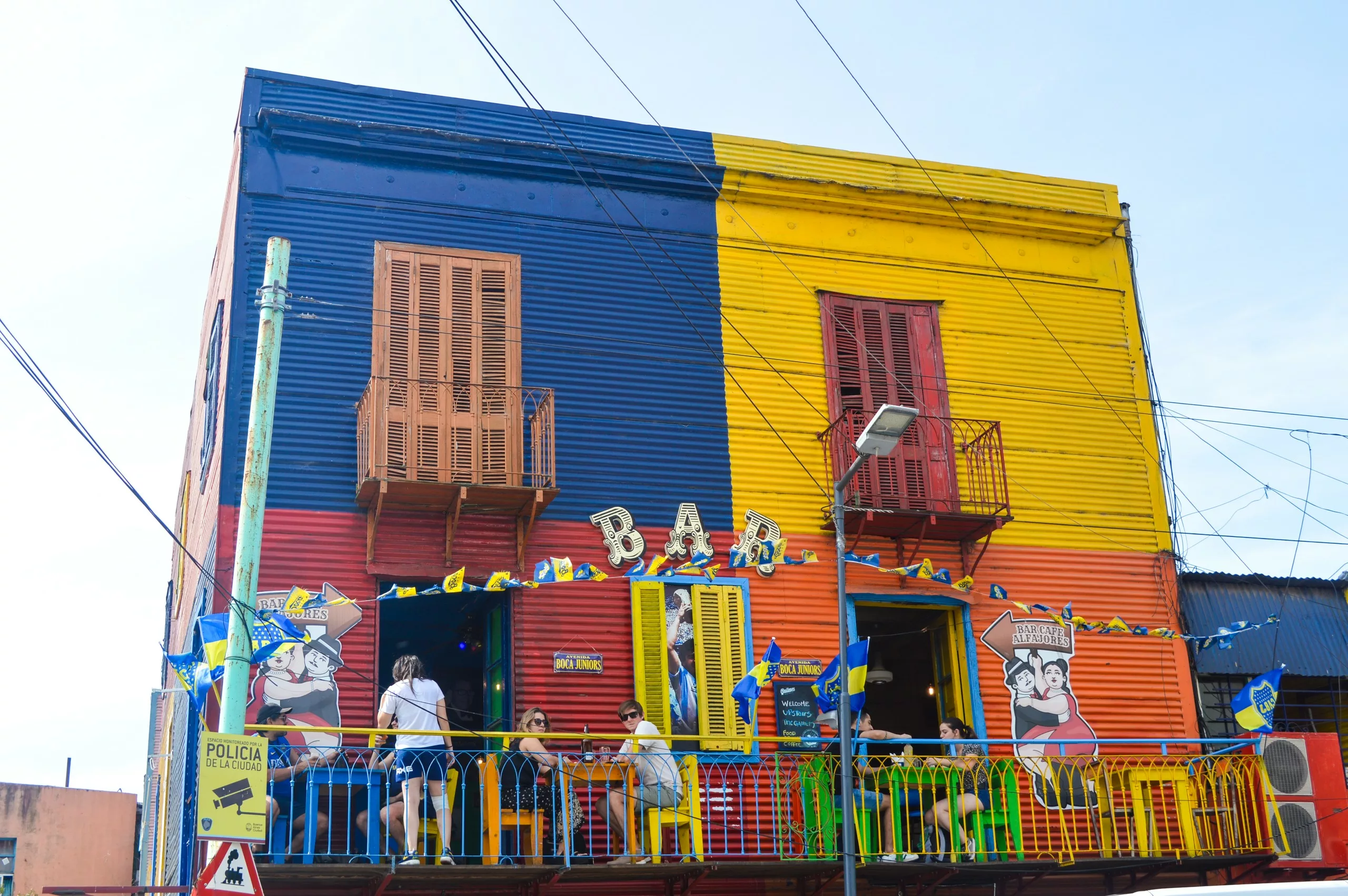 À Buenos Aires, un immeuble peint de couleurs vives, avec des panneaux jaunes, bleus et rouges, possède un balcon où des personnes sont assises sous un panneau "BAR". Des drapeaux sont accrochés à la balustrade ; des peintures murales représentant des visages