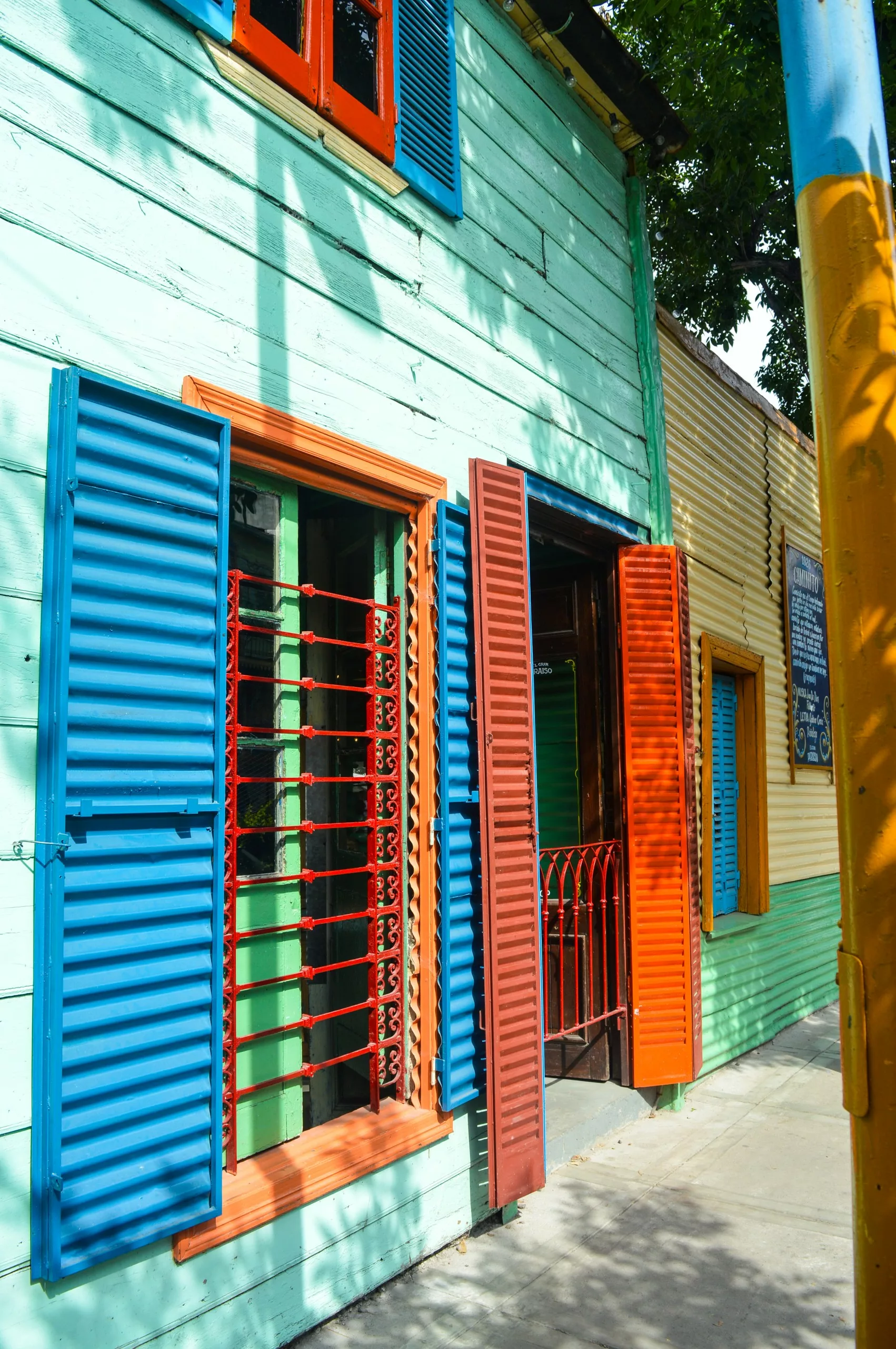 Un bâtiment avec des murs en bois vert pastel, des volets bleus et orange, des barreaux de fenêtres rouges et un poteau rayé jaune et bleu sur le trottoir