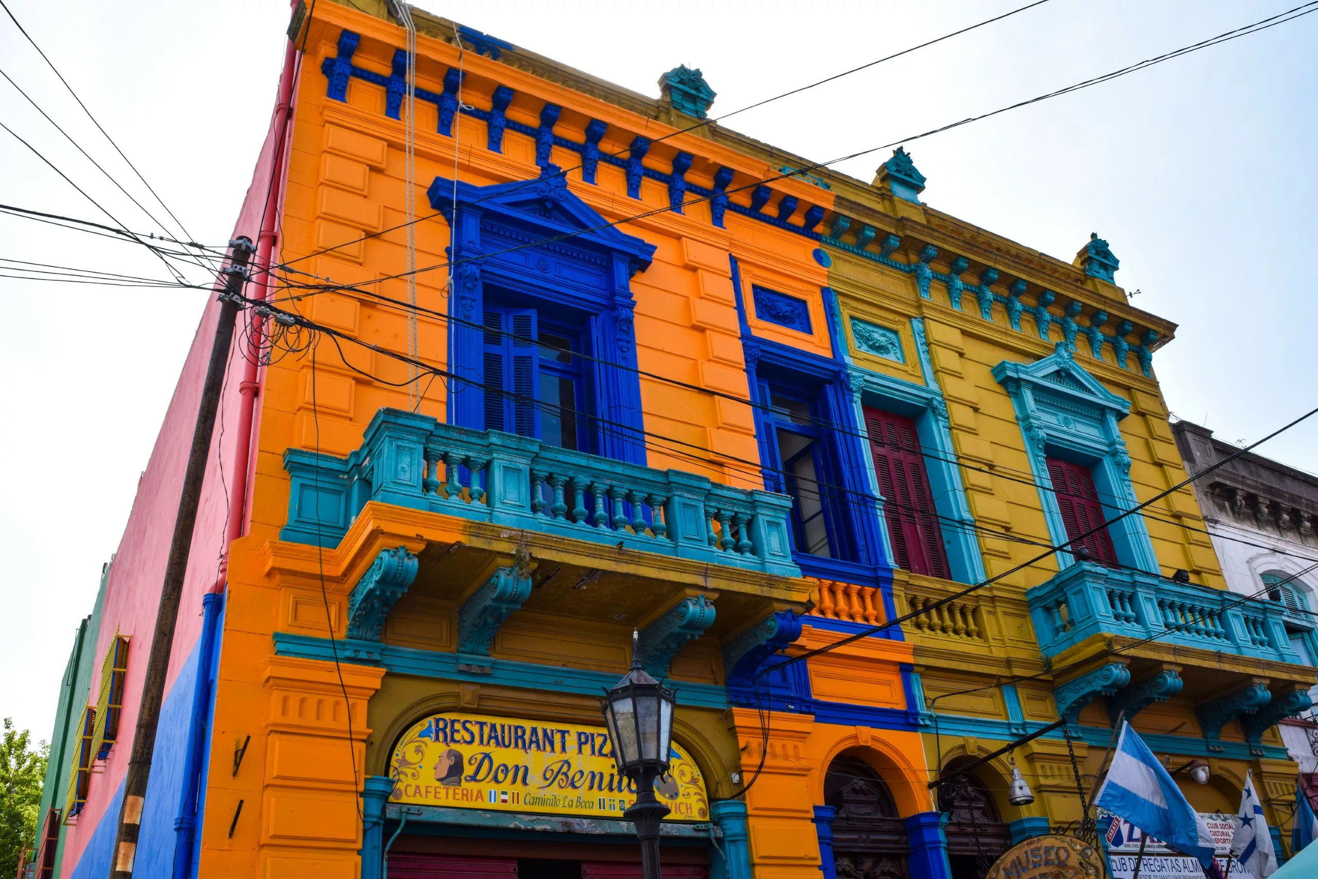 Un bâtiment de deux étages peint en orange, jaune et bleu vifs, avec des détails architecturaux ornés et des balcons. L'enseigne d'un restaurant pizzeria est visible en contrebas, et des fils électriques aériens passent devant.
