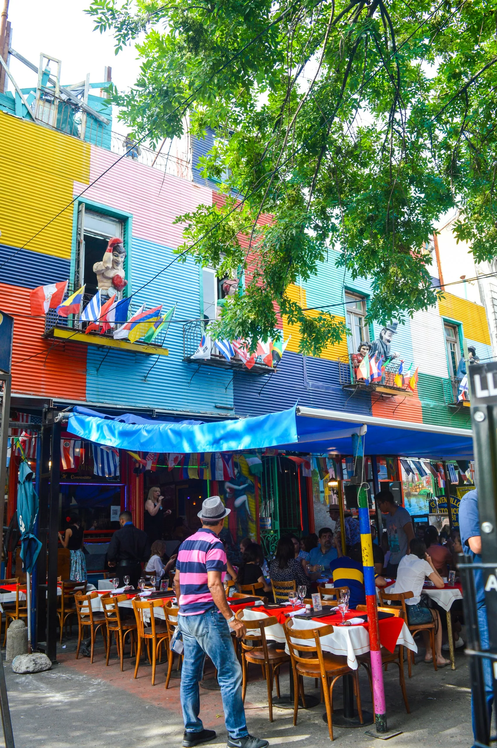 Un restaurant coloré avec des drapeaux en plein air dans une rue très animé de Buenos Aires. Des personnes sont attablées sous un auvent bleu.