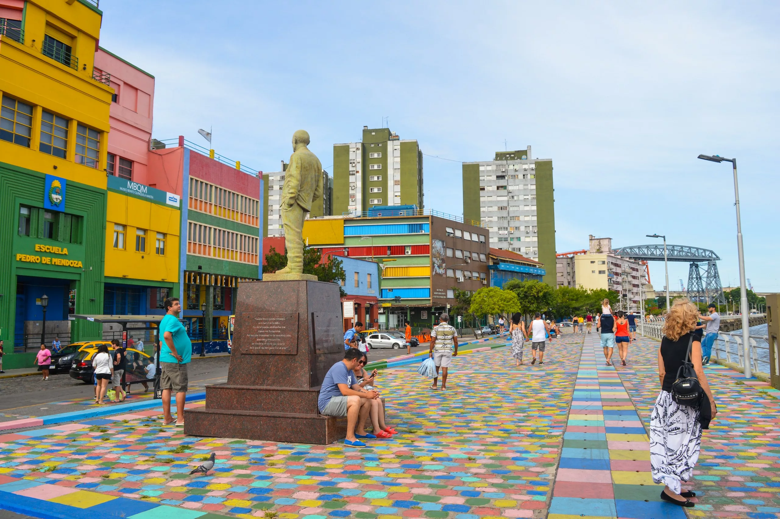 Des gens marchent le long d'un chemin coloré, pavé de mosaïques et bordé de bâtiments colorés. Une statue se dresse au centre.
