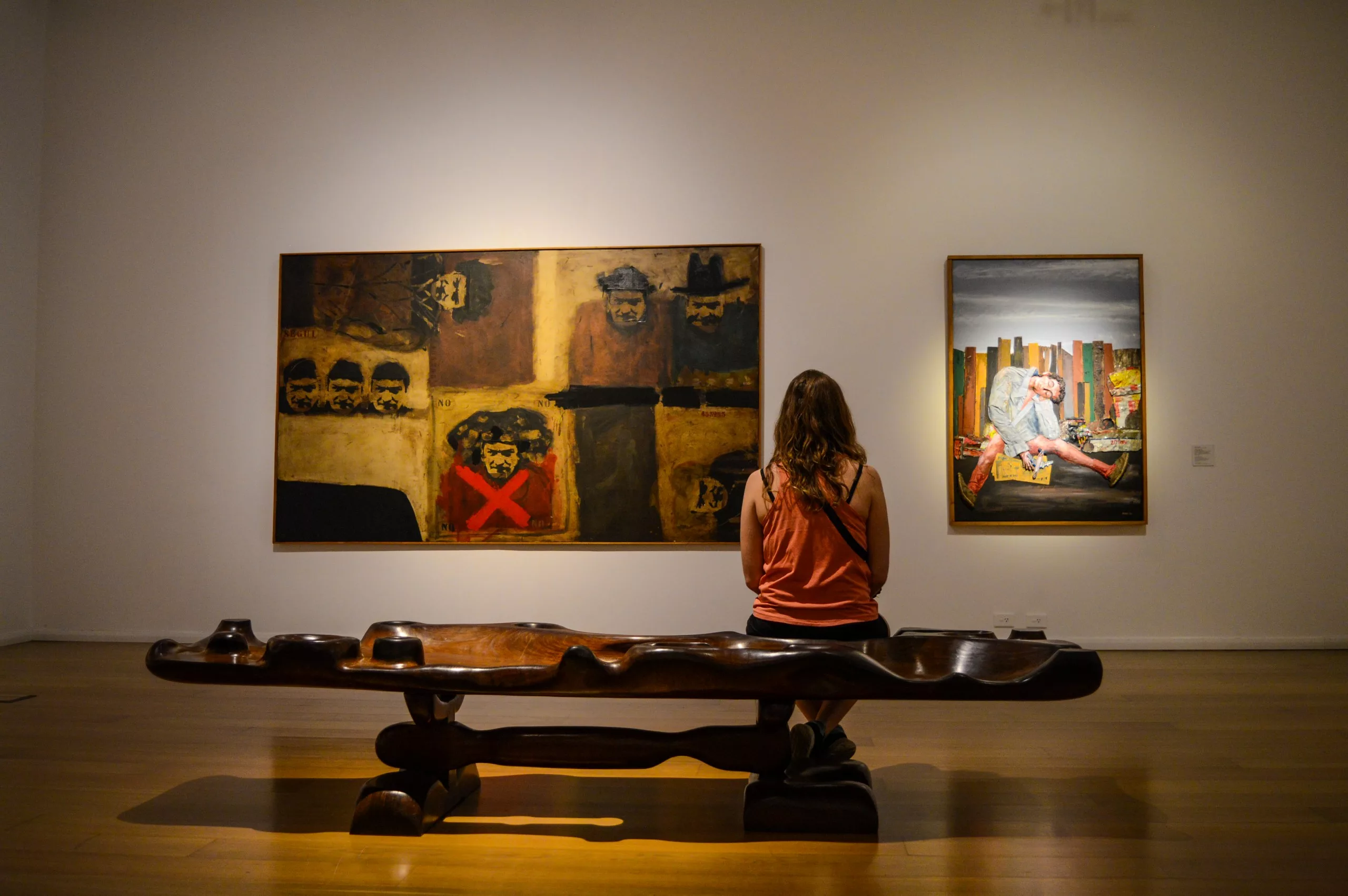 Une femme est assise sur un banc en bois dans un musée, profitant de l'ambiance calme. Des lumières douces mettent en valeur deux tableaux sur un mur blanc au-dessus d'un sol en bois poli.
