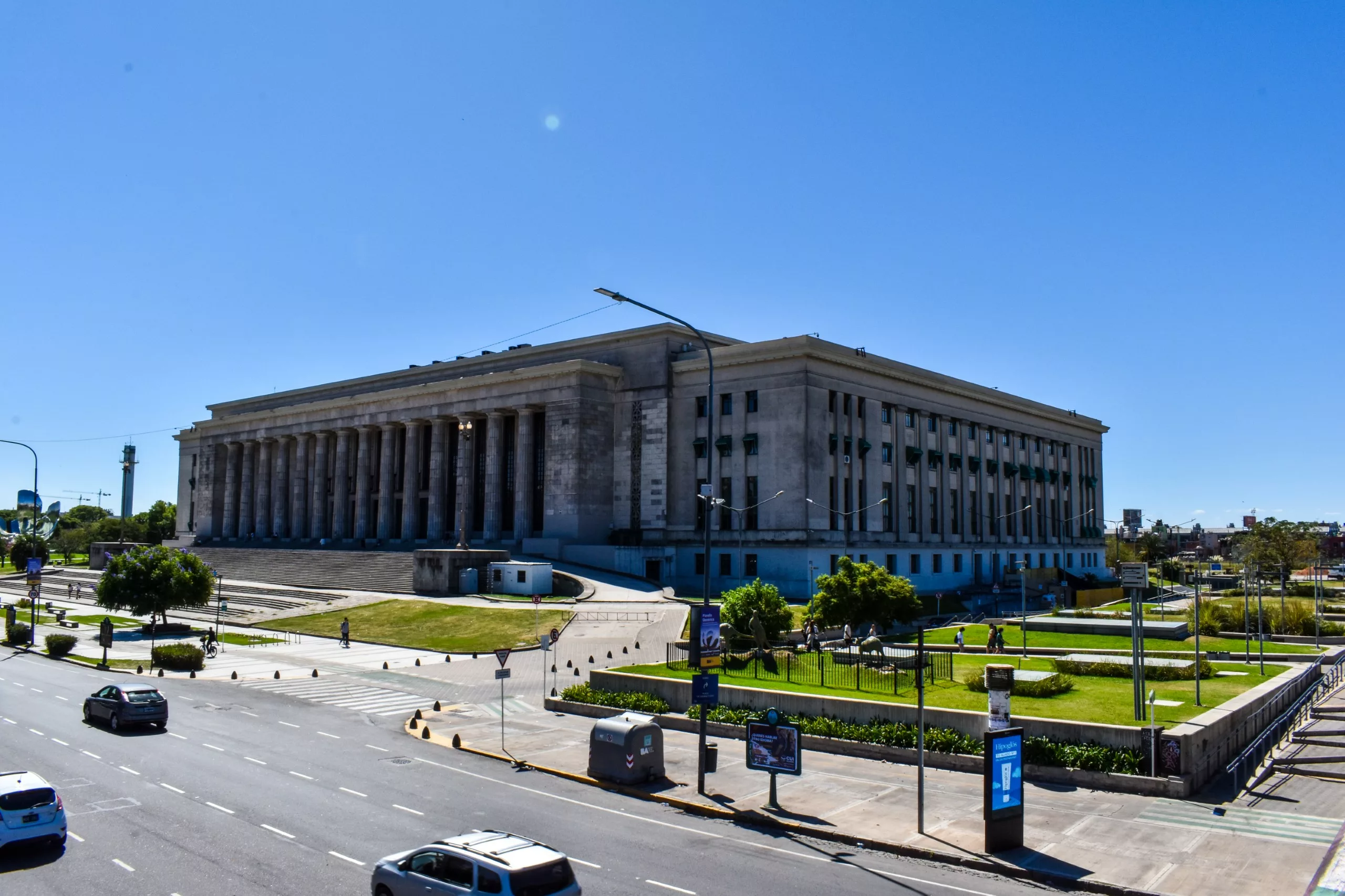 Un grand bâtiment néoclassique avec de hautes colonnes se dresse sur une plate-forme surélevée le long d'une rue animée de Buenos Aires. Les voitures passent devant des pelouses vertes, des marches mènent à l'entrée.