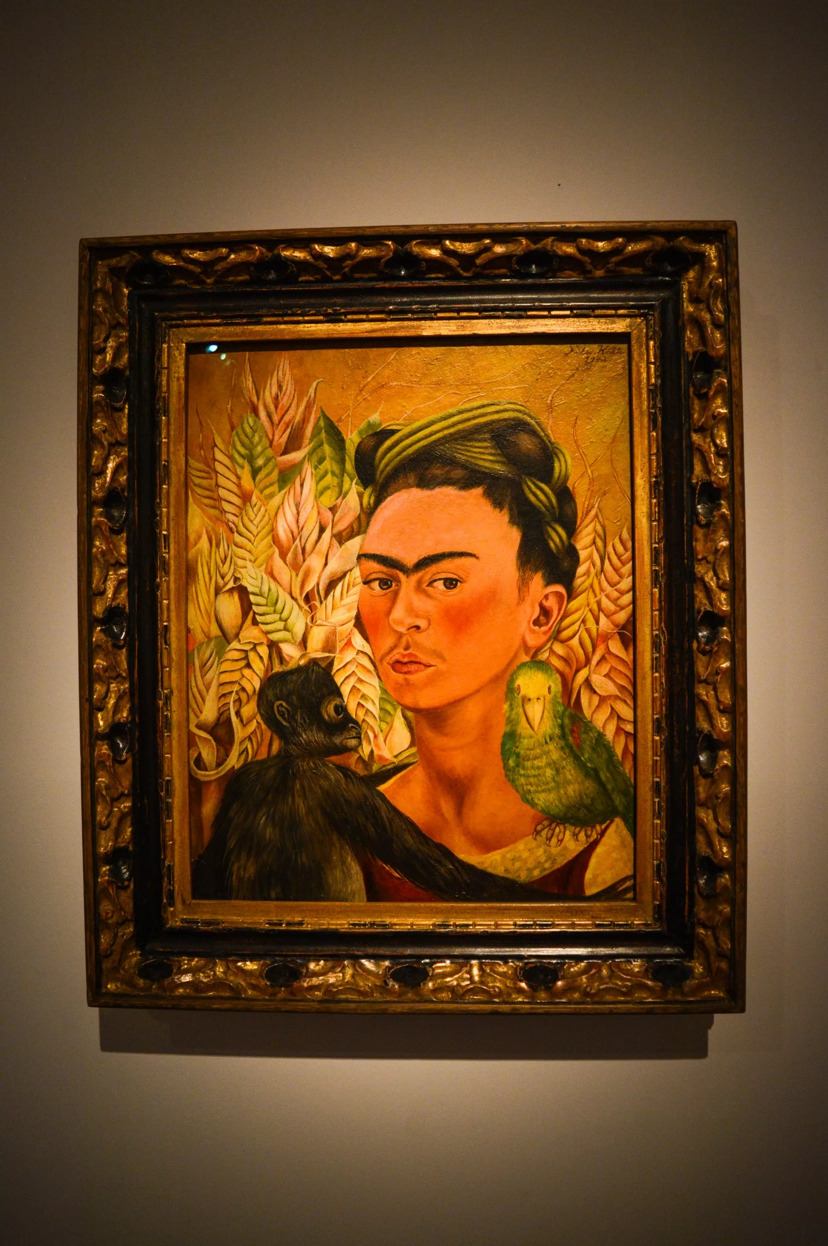Un tableau encadré représente une femme aux cheveux noirs, un singe sur son épaule et un perroquet vert à ses côtés sur fond de feuilles luxuriantes et de couleurs naturelles dans un cadre doré orné.