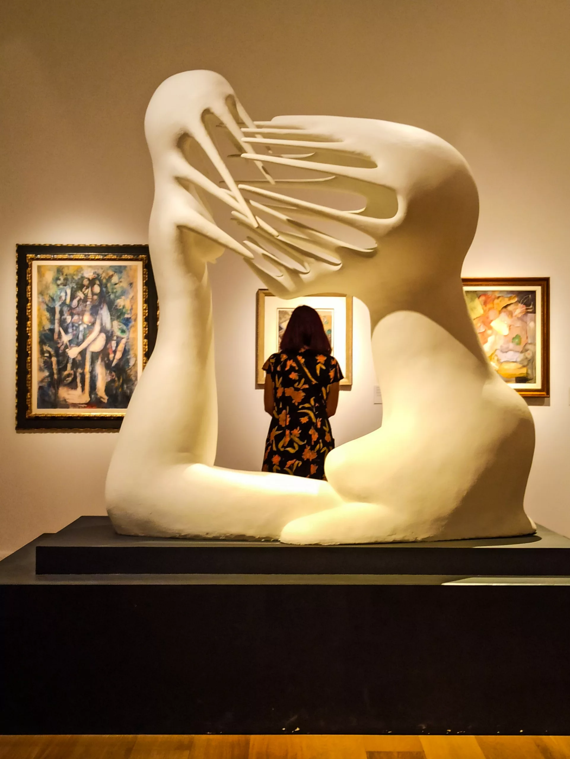 Une grande sculpture abstraite blanche, dont les formes ressemblent à des mains imbriquées, se trouve dans une galerie. Une visiteuse vêtue d'une robe à fleurs, regarde au loin, admirant des peintures encadrées sur le mur.