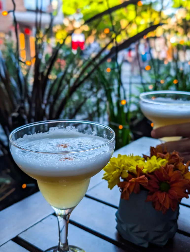 Deux cocktails jaunes mousseux reposent sur une table en bois à l'extérieur, à côté de fleurs vibrantes