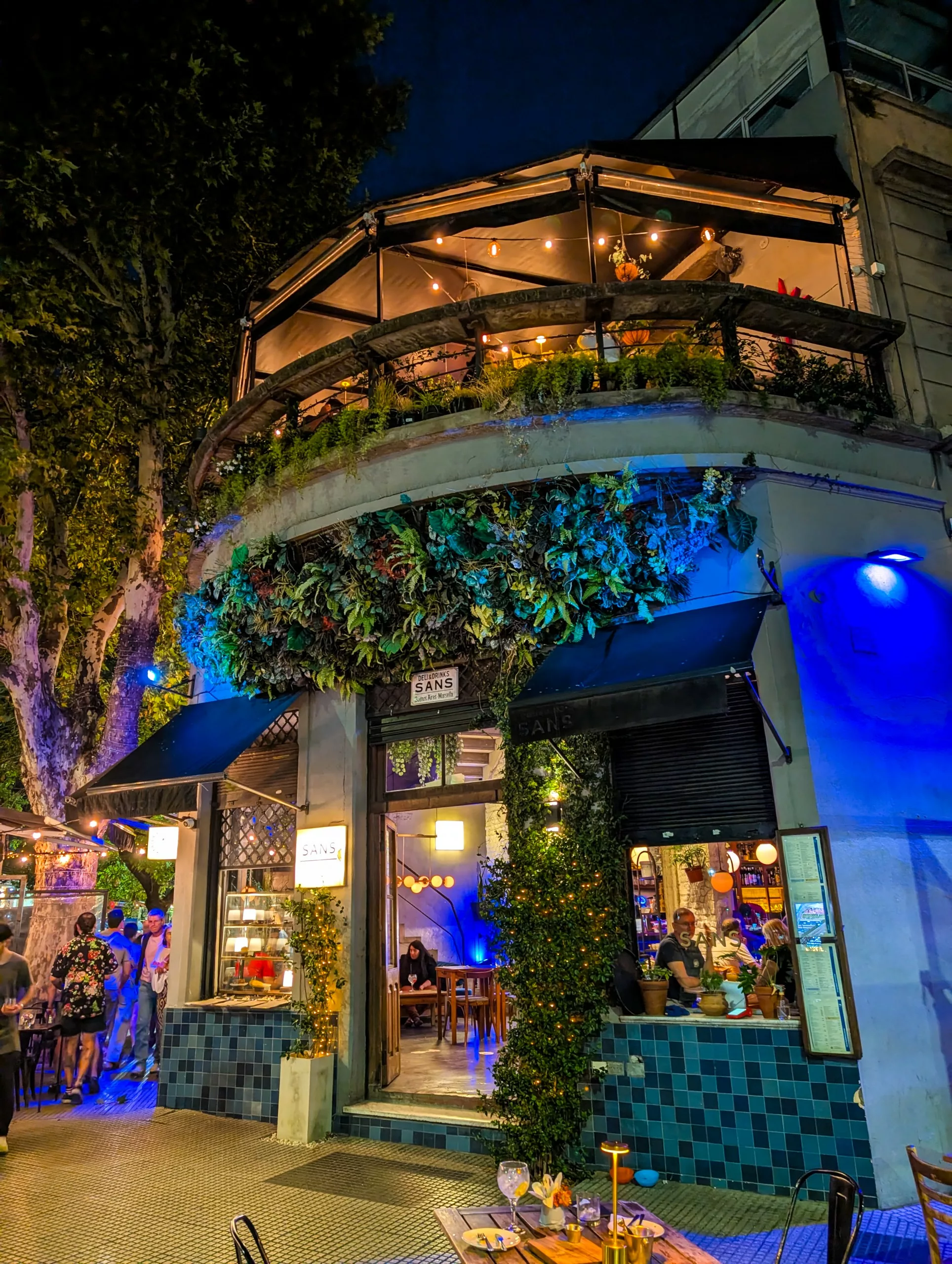 Un restaurant de deux étages à Buenos Aires avec un jardin vertical vert sur sa façade, des carreaux bleus à l'entrée et des places assises à l'extérieur. Des lumières chaudes illuminent les deux niveaux la nuit pendant que les gens dînent