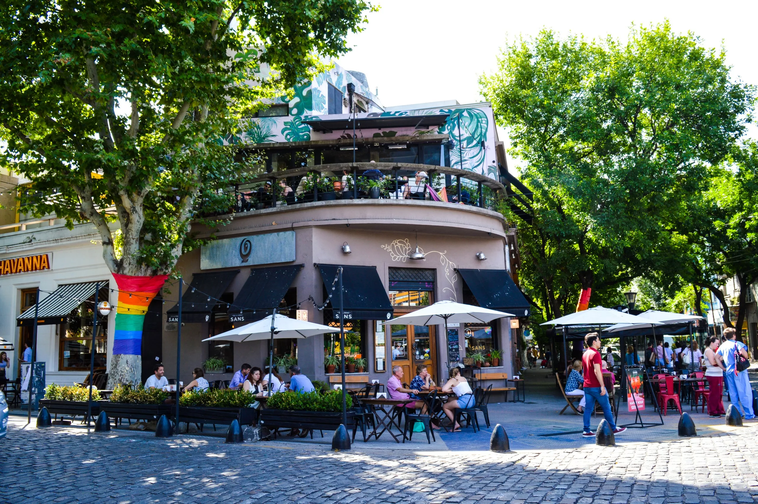 Un coin de rue avec un café et des sièges en plein air sous des parasols, des clients attablés, un drapeau de la fierté arc-en-ciel suspendu à un arbre