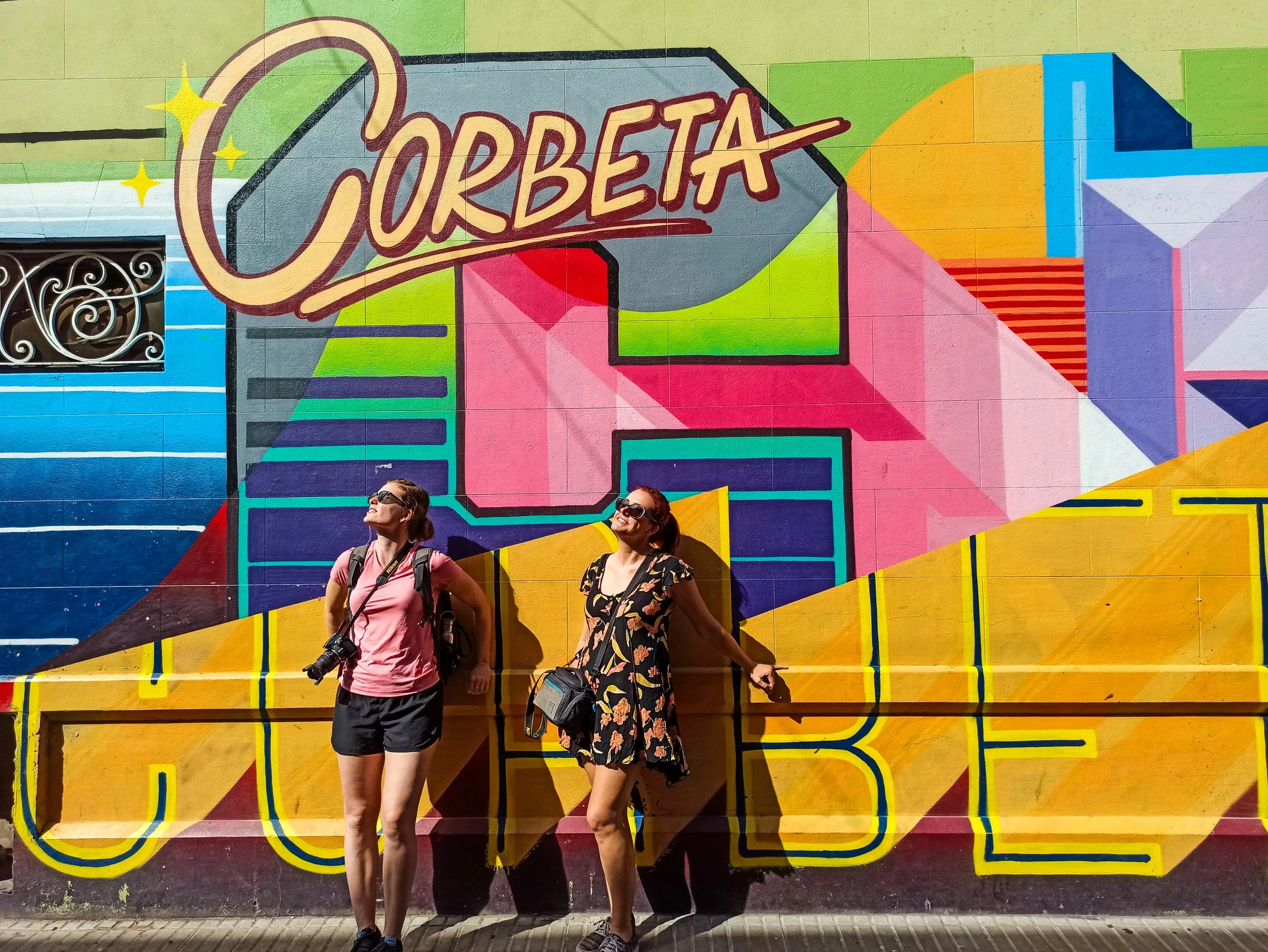 Deux personnes se tiennent devant une peinture murale colorée sur laquelle est inscrit "Corbeta", profitant de la lumière du soleil. Elles portent toutes deux un sac à bandoulière.