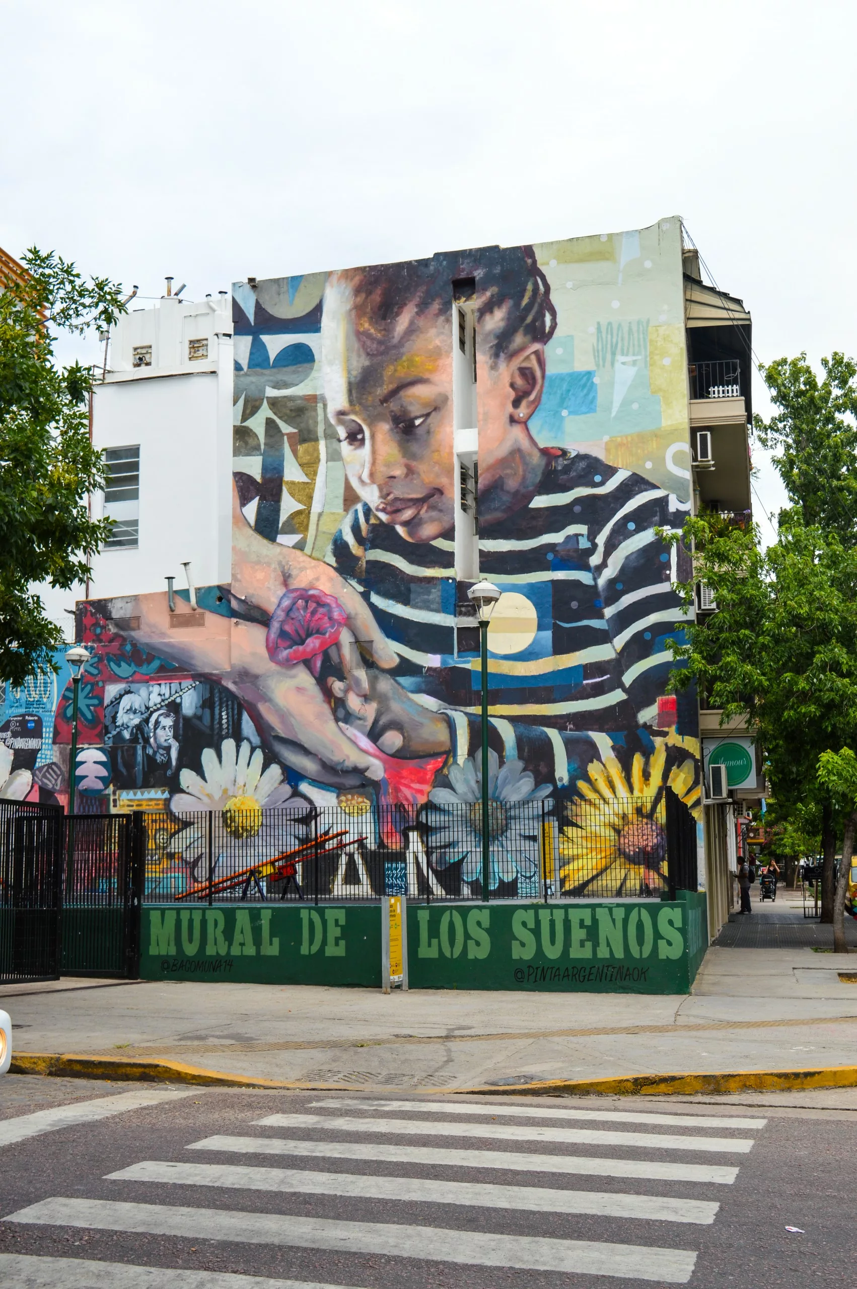 Une grande peinture murale sur un bâtiment du quartier Palermo à Buenos Aires montre un enfant tressé tenant une fleur rose. Les mains se tendent vers l'enfant, avec "Mural de los Sueños" en dessous.