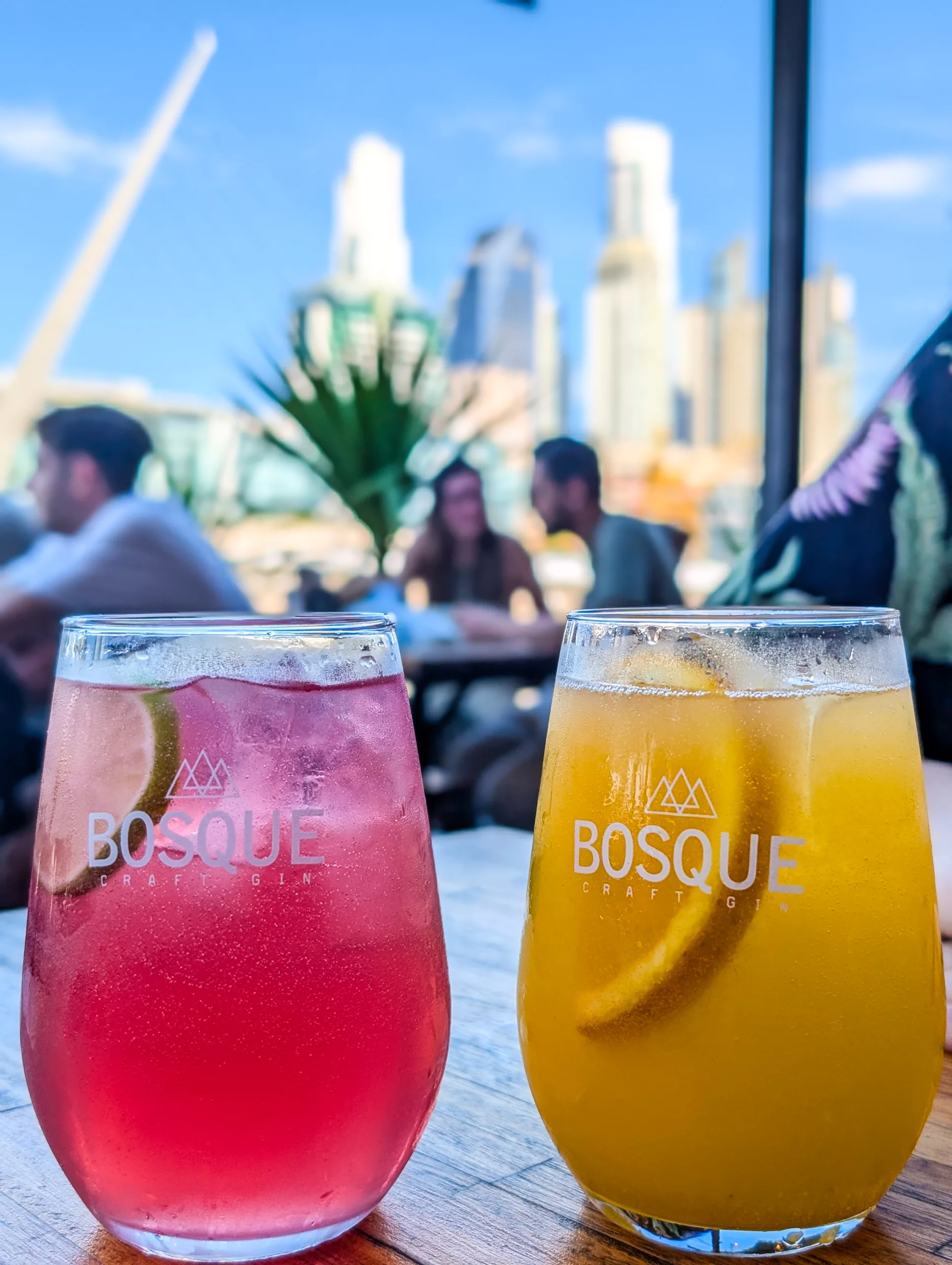 Deux verres de boissons colorées, étiquetées "Bosque Craft Gin", sont posés sur une table en bois. Les gens et les bâtiments de la ville se confondent à l'arrière-plan