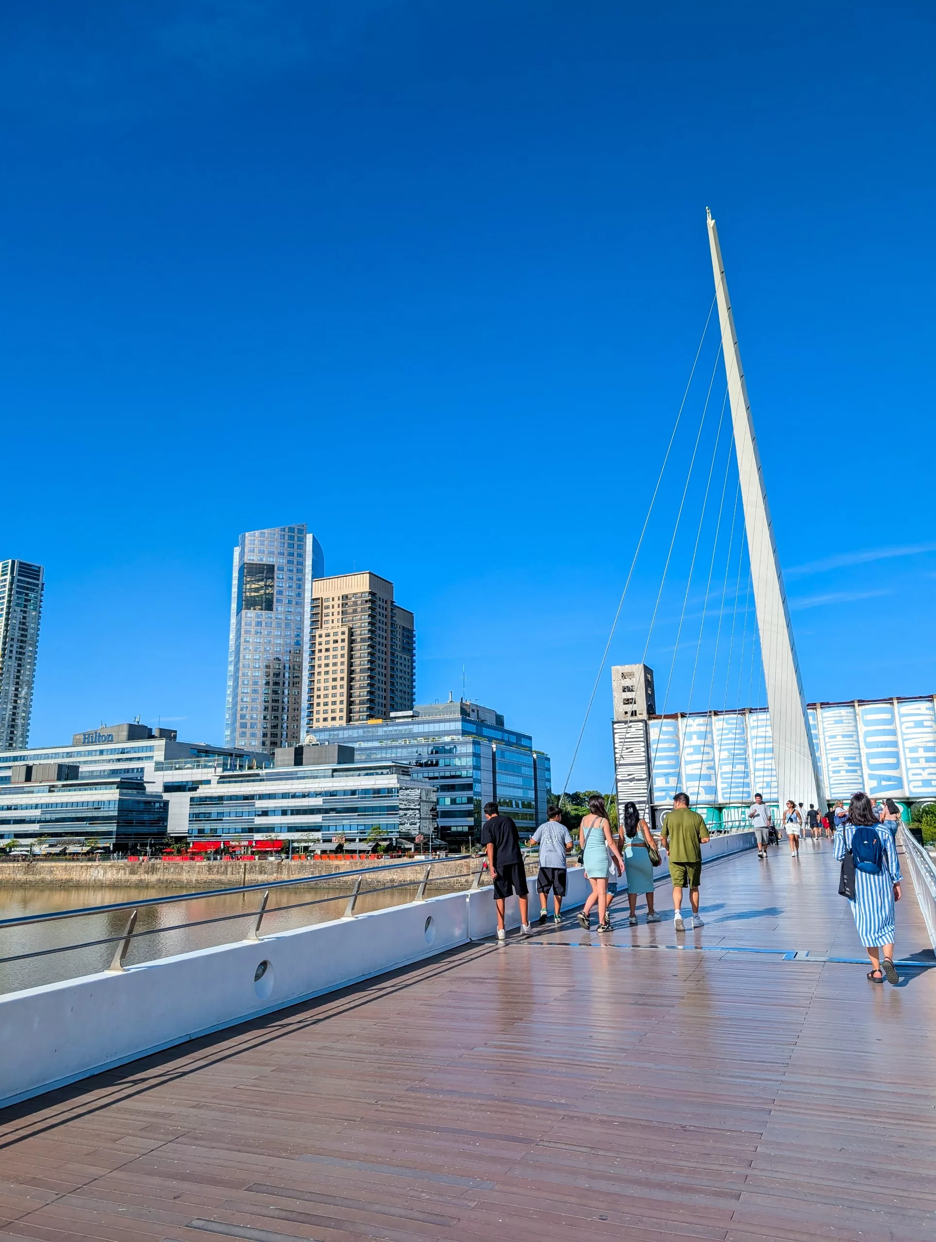 Des personnes marchent sur un pont piétonnier moderne à Buenos Aires, avec de hauts bâtiments en arrière-plan. Le pont se caractérise par un câble de soutien angulaire proéminent.