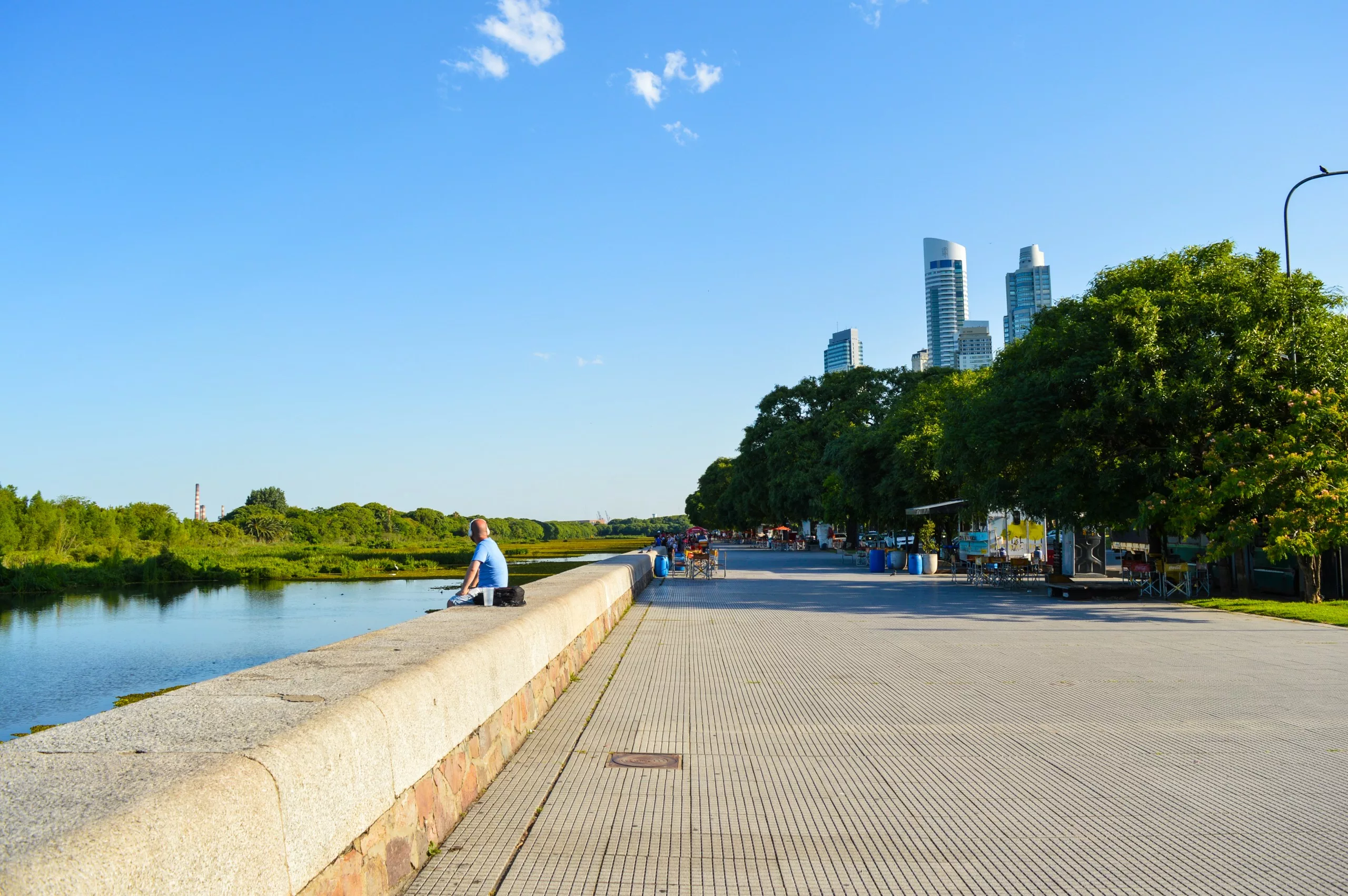 Un chemin pavé le long de la rivière avec une balustrade en pierre, avec une personne sur le rebord, des allées bordées d'arbres et des gratte-ciel modernes