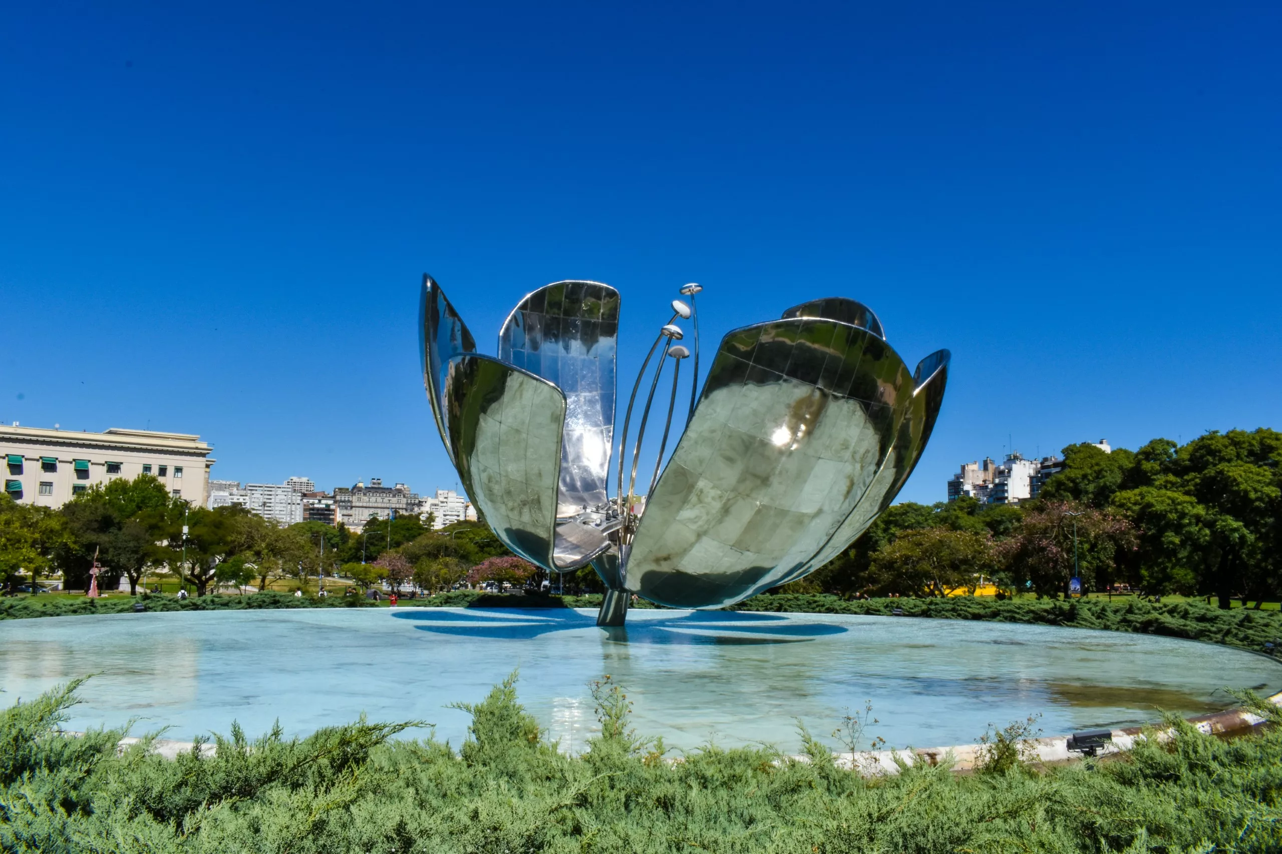 Une grande sculpture florale métallique aux pétales ouverts se dresse dans un bassin, entourée de verdure et de bâtiments urbains.