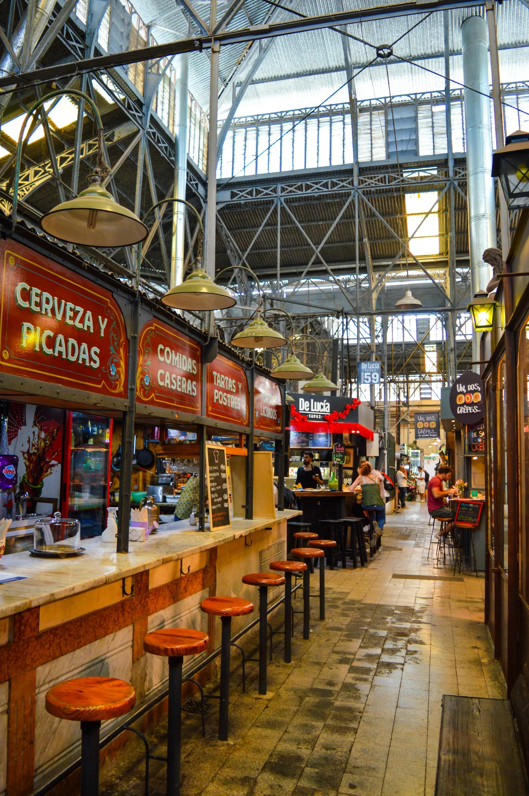 Une halle de marché avec de hauts plafonds métalliques, des stands de nourriture avec des panneaux en bois proposant de la bière, des snacks et des plats faits maison