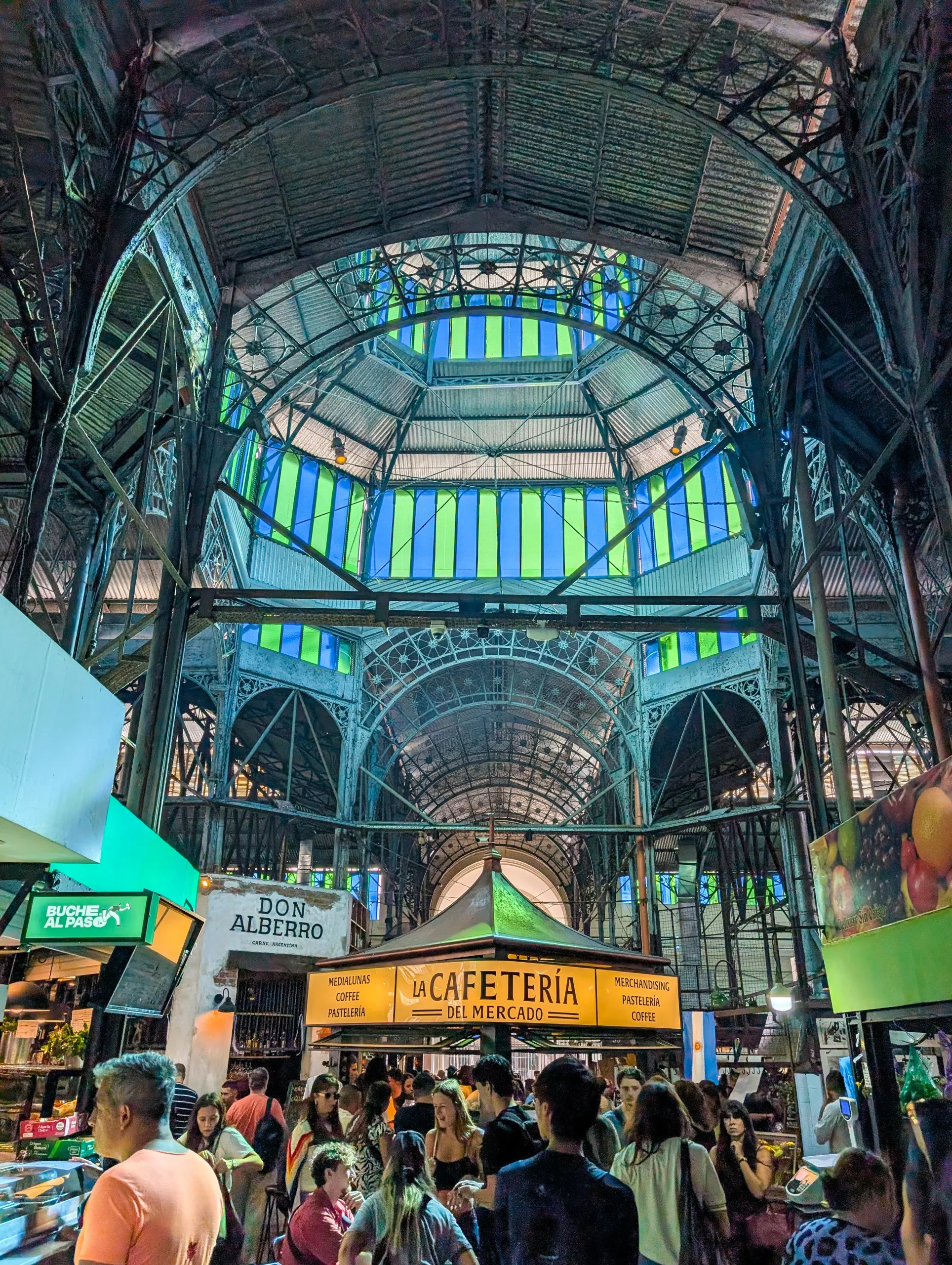 Les gens se rassemblent à l'intérieur d'un marché couvert animé à Buenos Aires, avec un haut plafond voûté et des vitraux colorés. Des enseignes de cafétérias et des étals animés remplissent la scène.