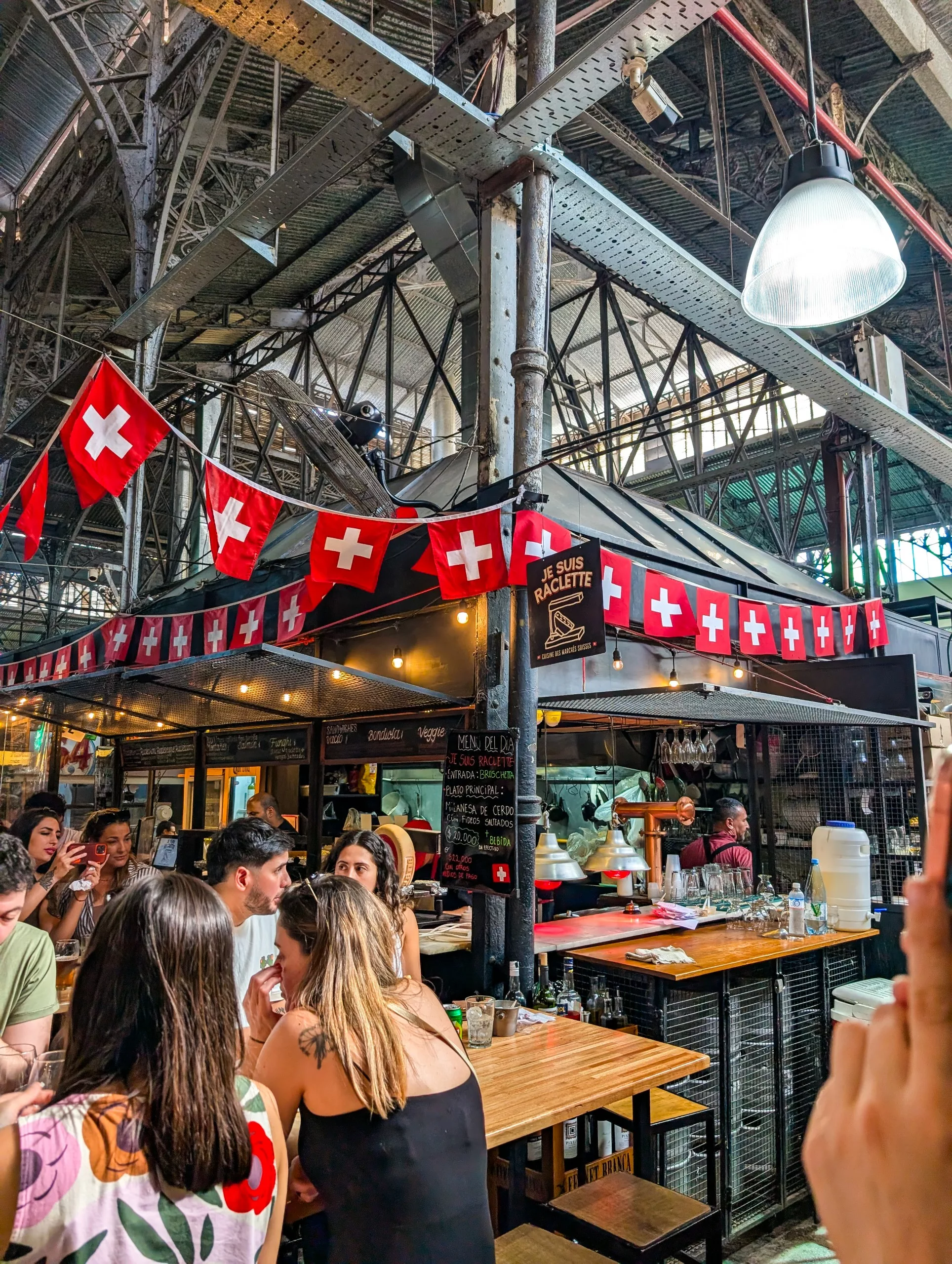 Scène de marché intérieur avec des gens assis à des tables en bois près d'un stand de nourriture décoré de drapeaux suisses. Des poutres et des éclairages industriels sont visibles au-dessus.