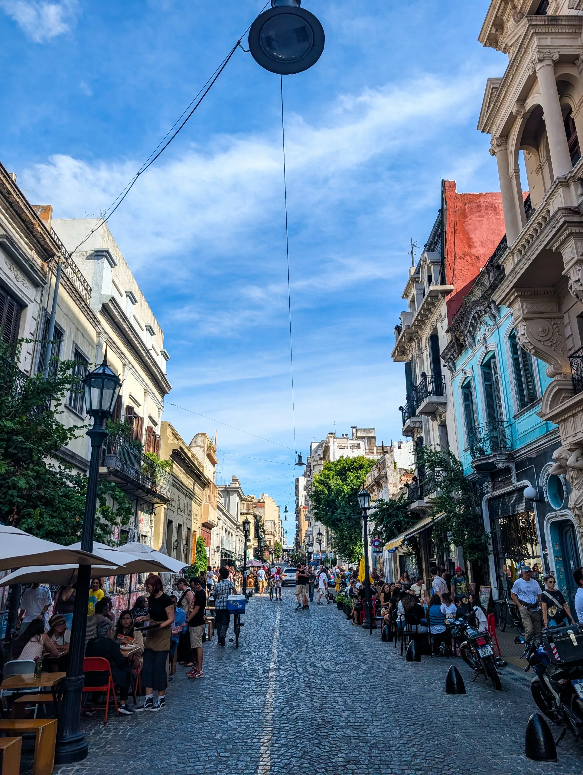 Une rue pavée de Buenos Aires, bordée de bâtiments historiques et de cafés en plein air, est animée par des gens qui marchent, s'assoient et se rencontrent sous un ciel bleu