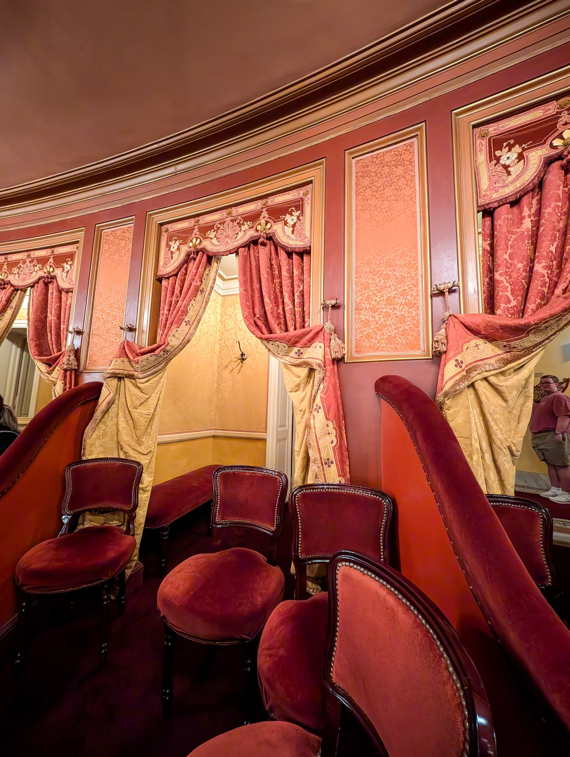 Une loge de théâtre avec des chaises en velours rouge et des rideaux ornés d'or et de rouge, offrant un design opulent et classique avec des couleurs riches, des panneaux muraux décoratifs et des motifs détaillés.