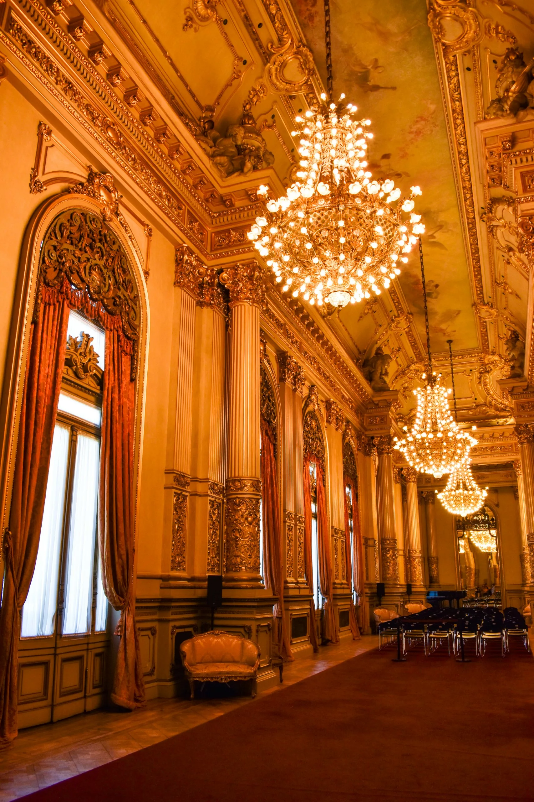 Hall de théâtre orné avec de hautes fenêtres cintrées drapées de rideaux rouges, des lustres en cristal, des détails dorés sur les murs et les colonnes, et des chaises disposées à l'extrémité.