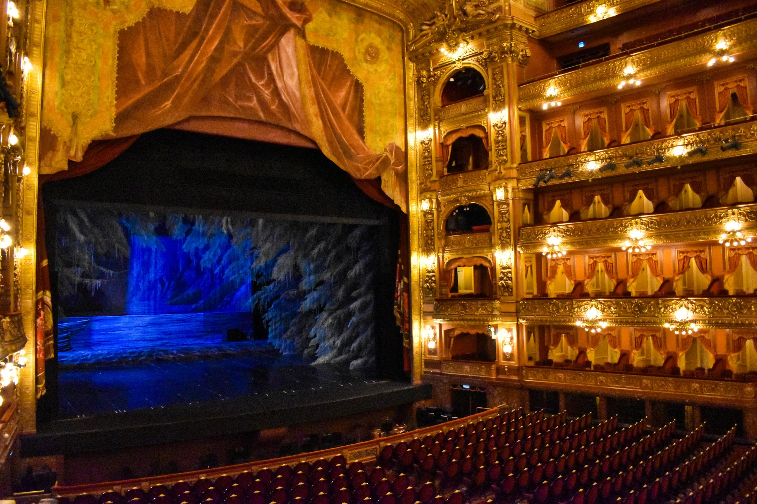 Intérieur de théâtre doré, à visiter à Buenos Aires, avec des rangées de sièges rouges face à une scène où se trouve un décor sombre. Trois niveaux de loges sont alignés sur le côté droit ; la toile de fond présente une scène abstraite bleue et grise.