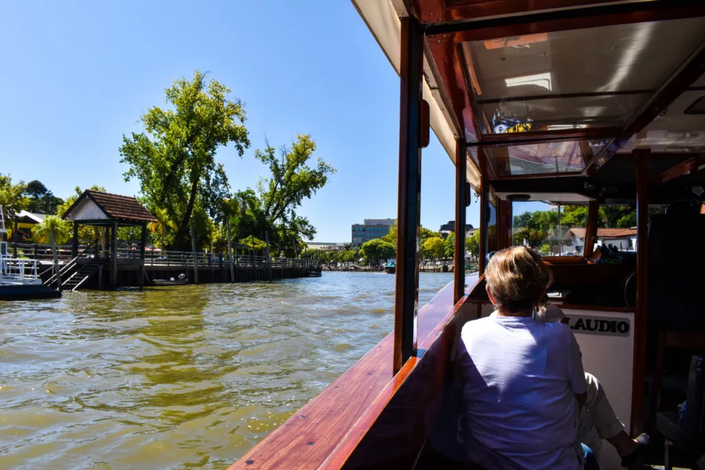 Une personne aux cheveux courts est assise sur un bateau en bois, regardant une vue de la rivière avec des arbres, des quais et des bâtiments