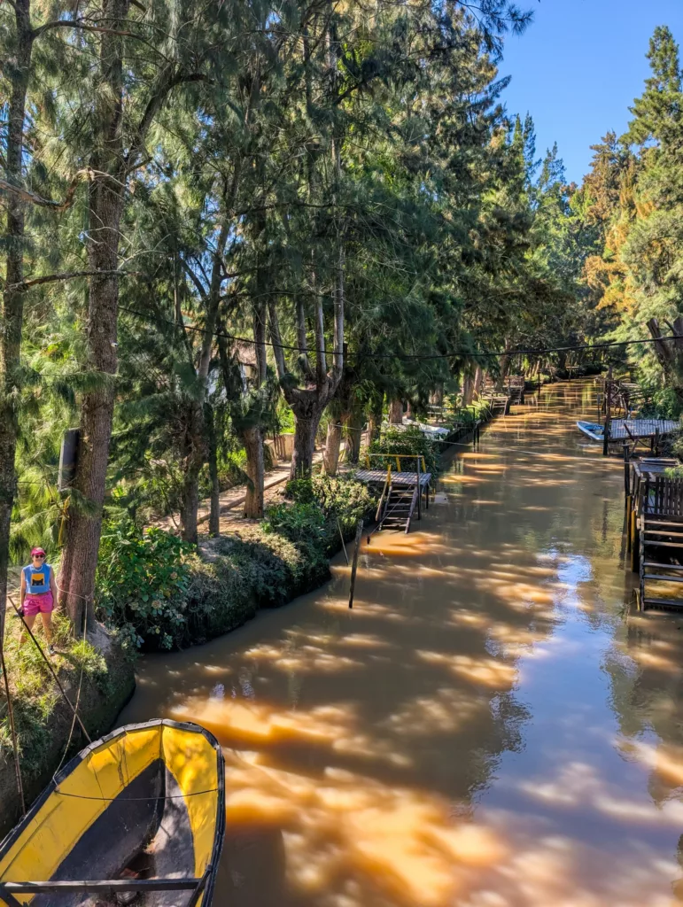Une rivière étroite et boueuse, bordée d'arbres et de quais en bois de part et d'autre. Un bateau jaune est amarré à proximité et une personne se tient au bord de l'eau.
