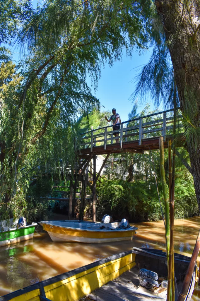 Une personne se tient sur une passerelle en bois au-dessus d'une rivière brune. Plusieurs bateaux sont amarrés près de la rive, des arbres verdoyants entourent la zone
