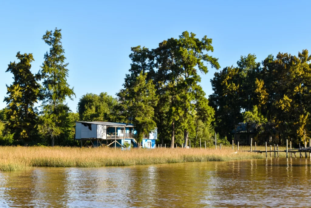 Une maison sur pilotis se dresse parmi de grands arbres et de l'herbe au bord d'une rivière calme sous un ciel bleu clair.