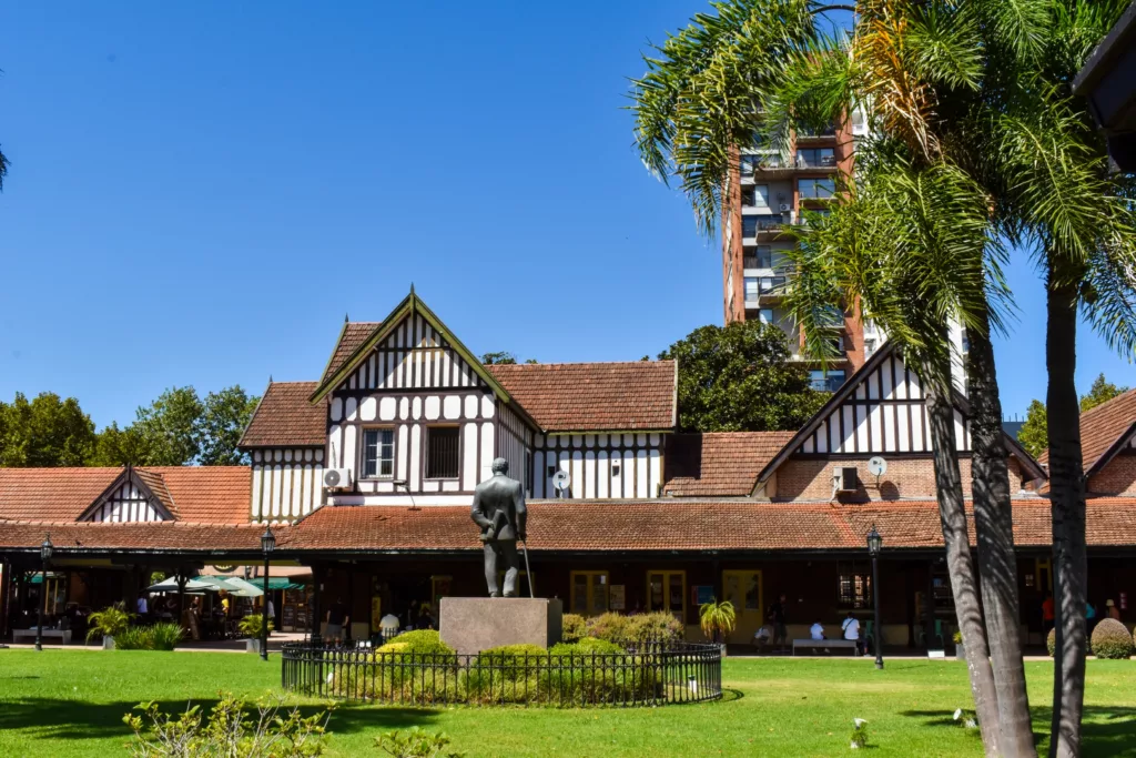 Une statue se dresse sur un piédestal dans une zone herbeuse devant un bâtiment de style Tudor encadré par des palmiers et une architecture moderne à l'arrière-plan.