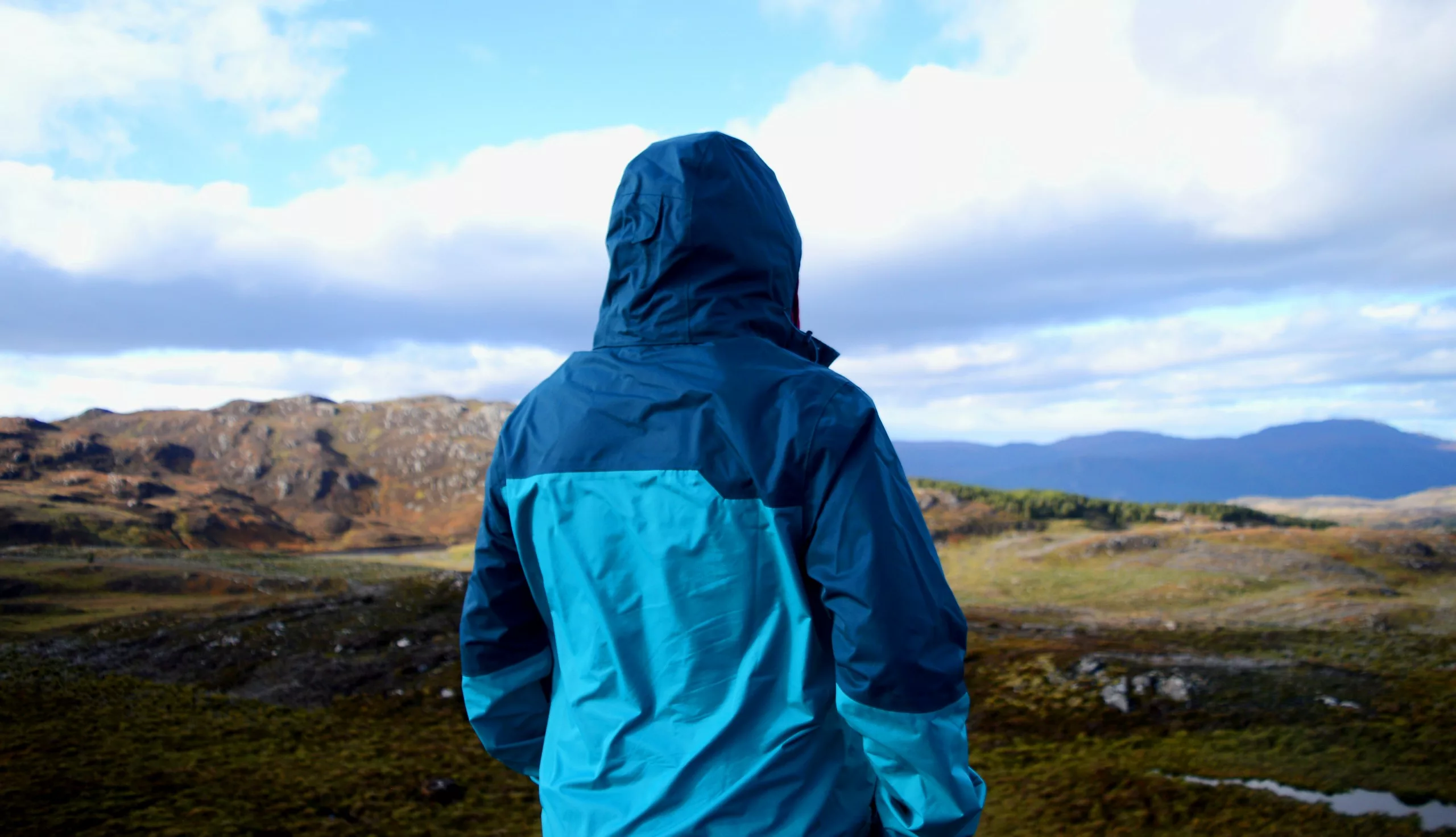 Une personne portant un imperméable à capuche bleue se tient à l'extérieur, face à un paysage de collines aux couleurs vertes et marrons.