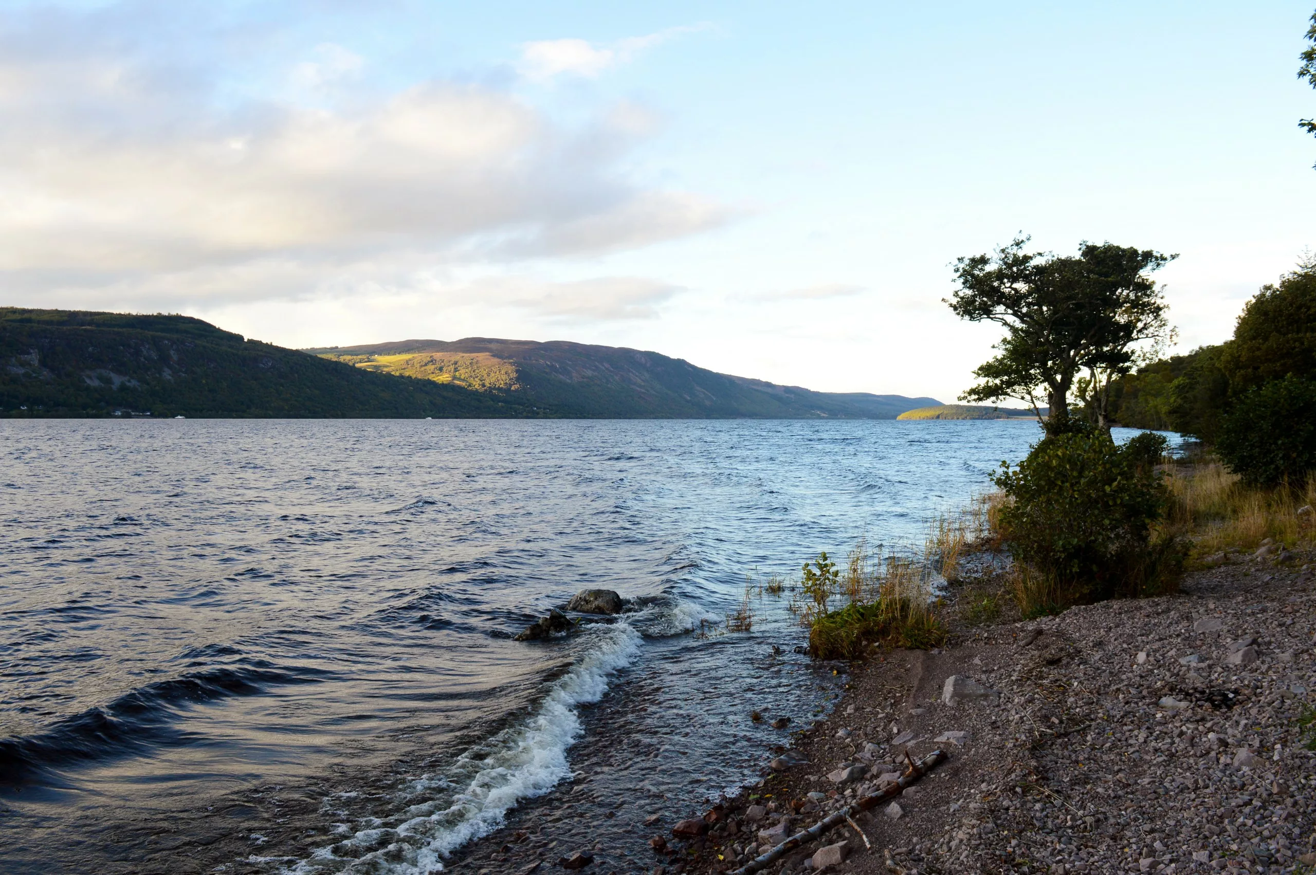 Visiter le loch ness : un grand lac entouré de collines, avec des vagues frappant doucement un rivage rocheux et quelques arbres sur la droite