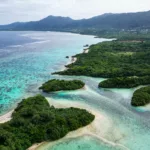Une vue aérienne d'un littoral aux eaux turquoise, parsemé de petites îles verdoyantes et d'une végétation luxuriante, avec des montagnes en arrière