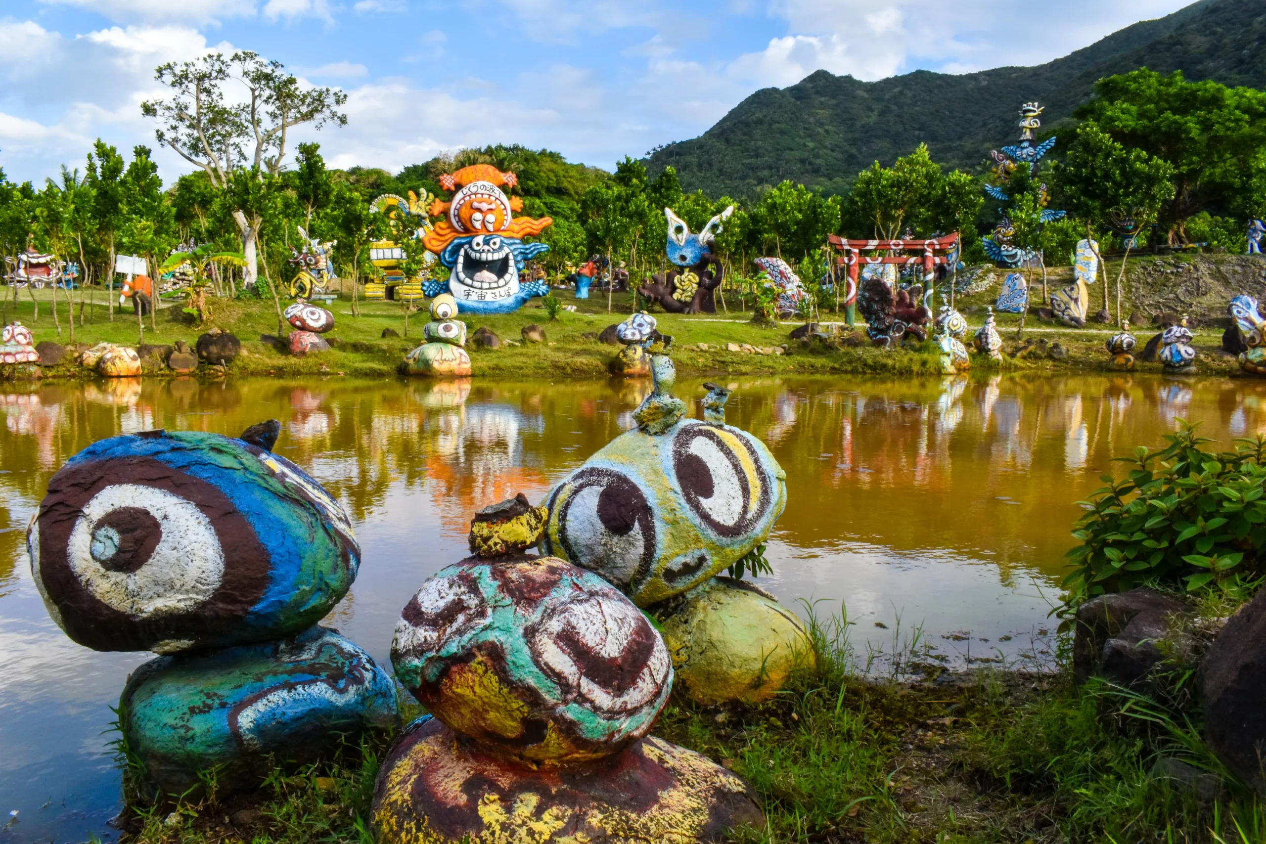 Des sculptures en pierre peintes, ornées de motifs colorés et tourbillonnants ainsi que de visages d'animaux, sont disposées au bord d'un étang
