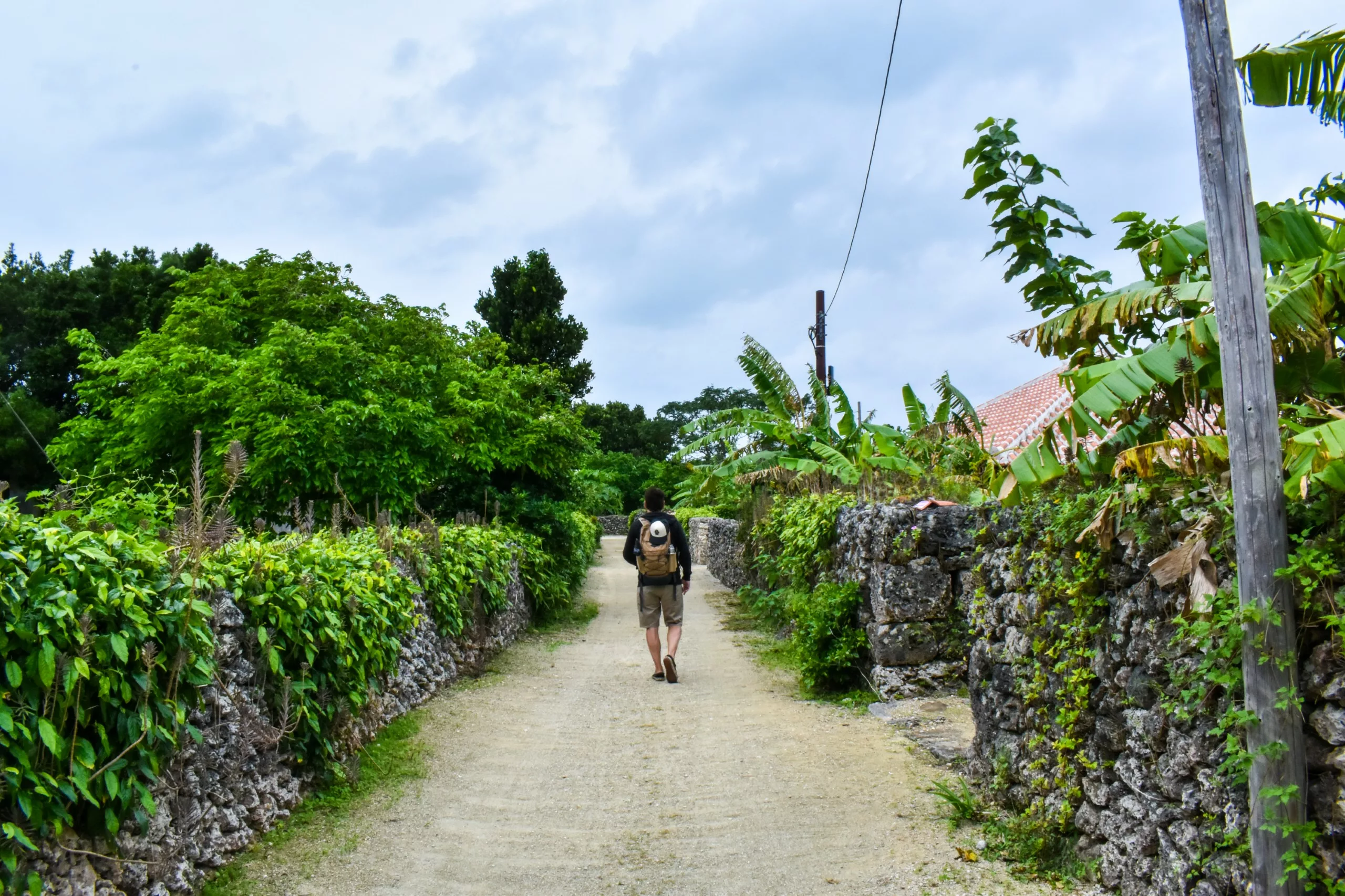 Un visiteur marche avec son sac à dos sur un chemin de terre bordé de murs de pierre et de végétation, sous un ciel nuageux. Des arbres et des toits se dessinent à l'arrière-plan.