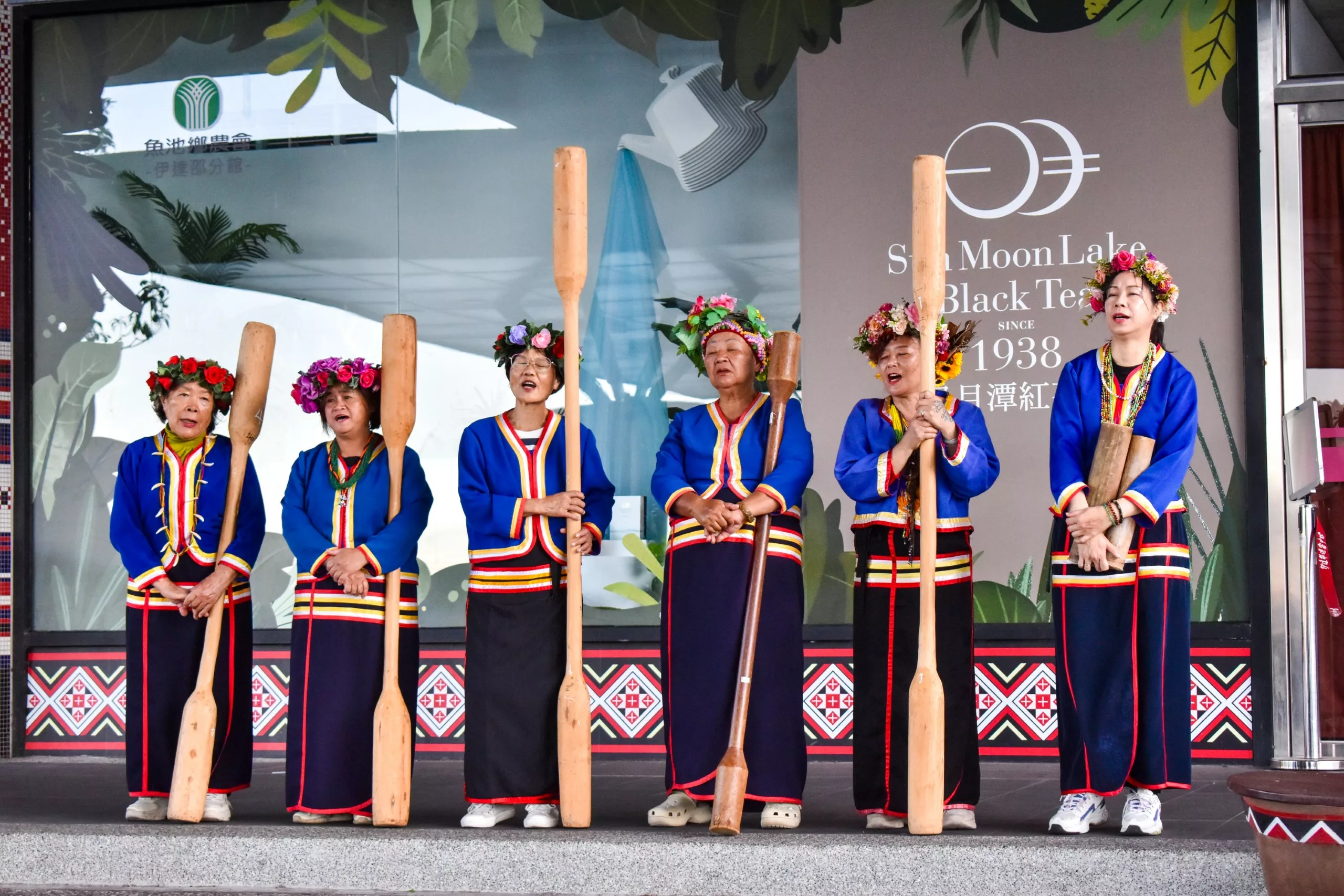 Six femmes portant des vêtements traditionnels et des coiffes fleuries se tiennent en rang, tenant de grandes pagaies en bois devant un bâtiment portant des panneaux "Sun Moon Lake Black Tea" (thé noir du lac Sun Moon) et des feuillages décoratifs.