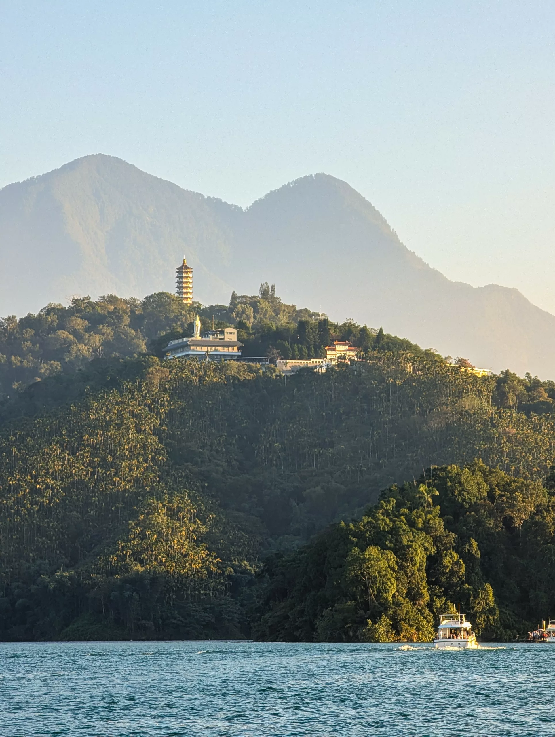 Vue d'une colline boisée avec une pagode et des bâtiments, sur fond de montagnes. Le lac Sun Moon brille au premier plan, où deux bateaux attendent le prochain visiteur sous la lumière du soleil de fin d'après-midi.