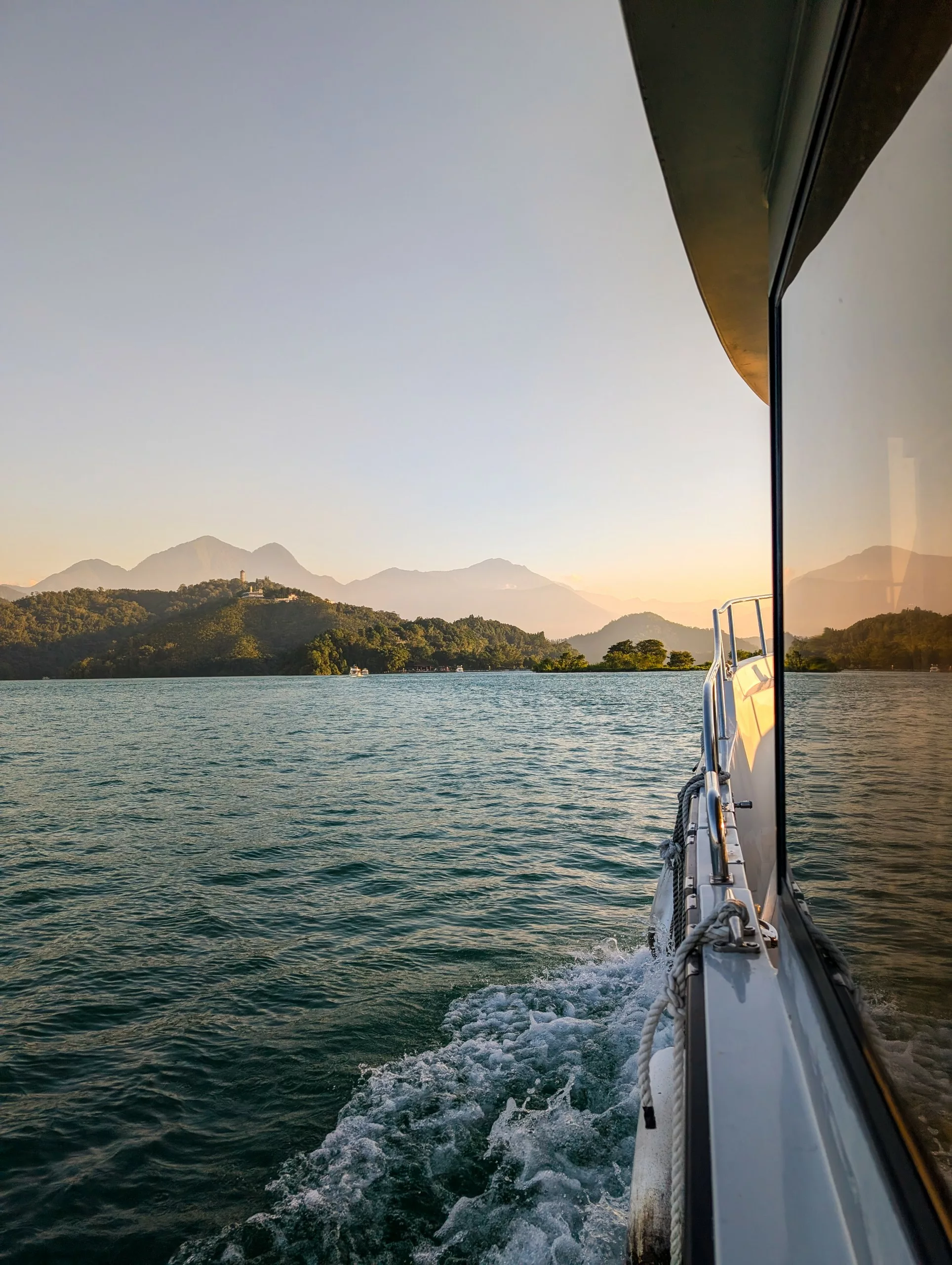 Un bateau navigue sur le lac Sun Moon, avec des montagnes et des arbres au loin. La lumière chaude du coucher de soleil se reflète sur les fenêtres.