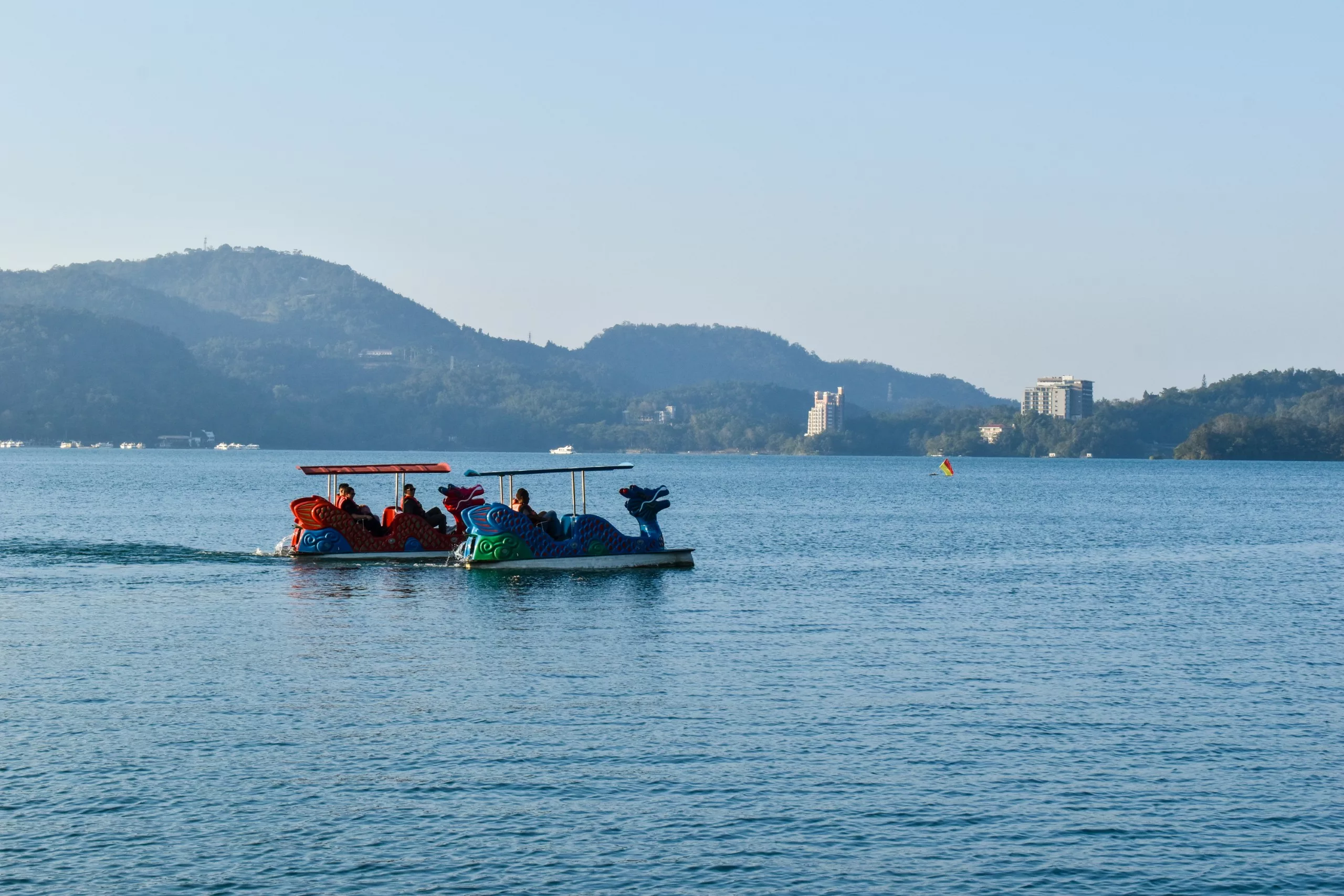 Deux pédalos colorés en forme de dragon transportent plusieurs personnes sur un lac calme au lac Sun Moon, avec des collines, des bâtiments et un ciel clair en arrière-plan, invitant tous les visiteurs à profiter des vues panoramiques.