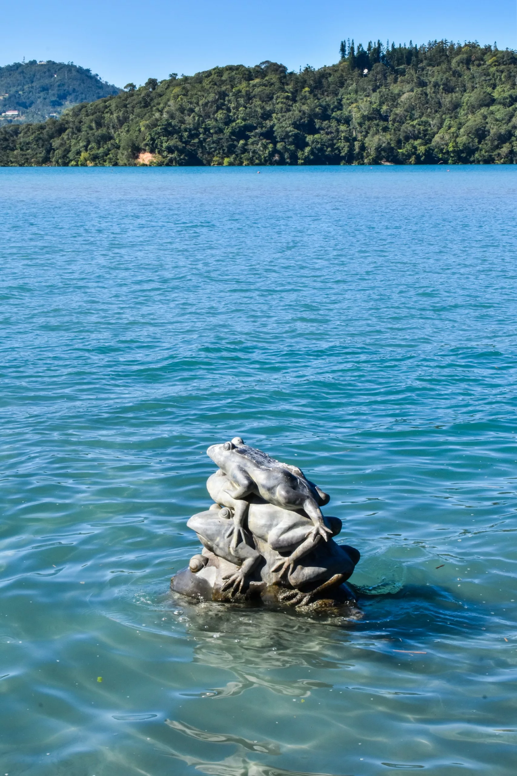 Une sculpture en pierre représentant plusieurs grenouilles empilées les unes sur les autres émerge de l'eau du lac Sun Moon, avec un rivage boisé et des collines visibles à l'arrière-plan.