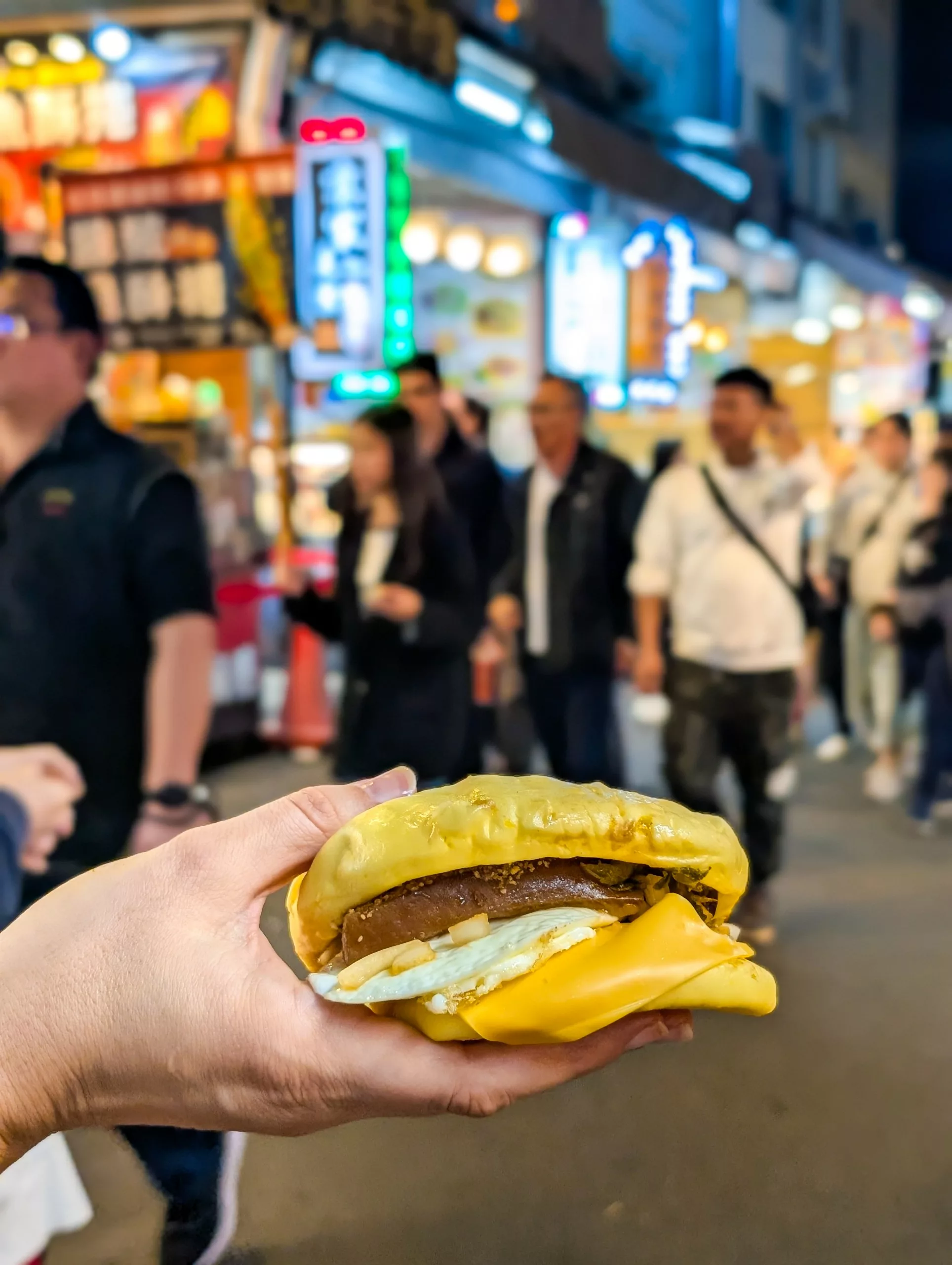 Une main tient un bao, pain brioché chinois, avec du fromage, un œuf et de la viande sur un marché nocturne en plein air très fréquenté.
